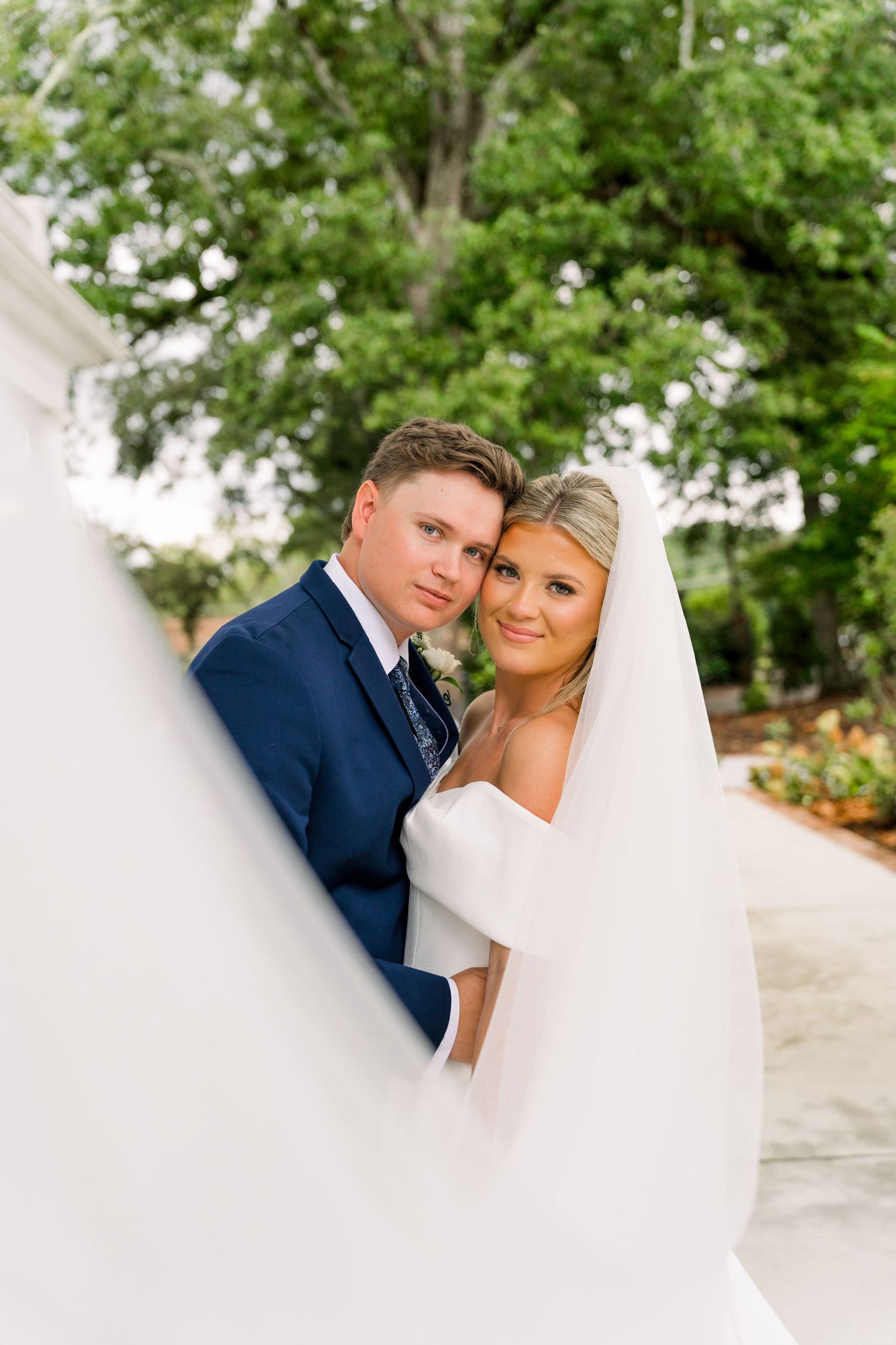 Bride in white gown and veil poses with groom in navy suit against white column, lush green trees in background.