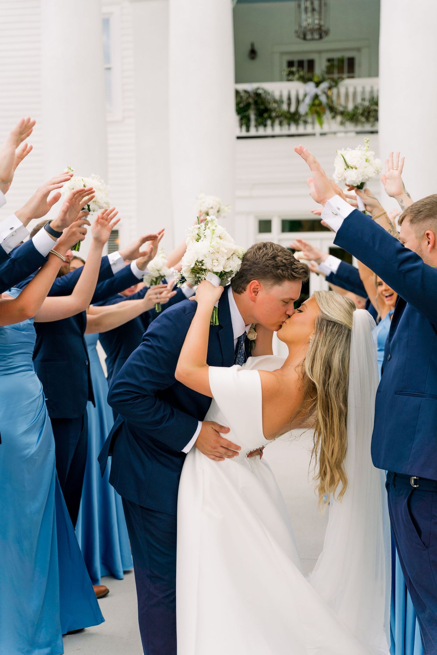 Bride and groom share a kiss while wedding party in blue raises arms in celebration outside white venue.