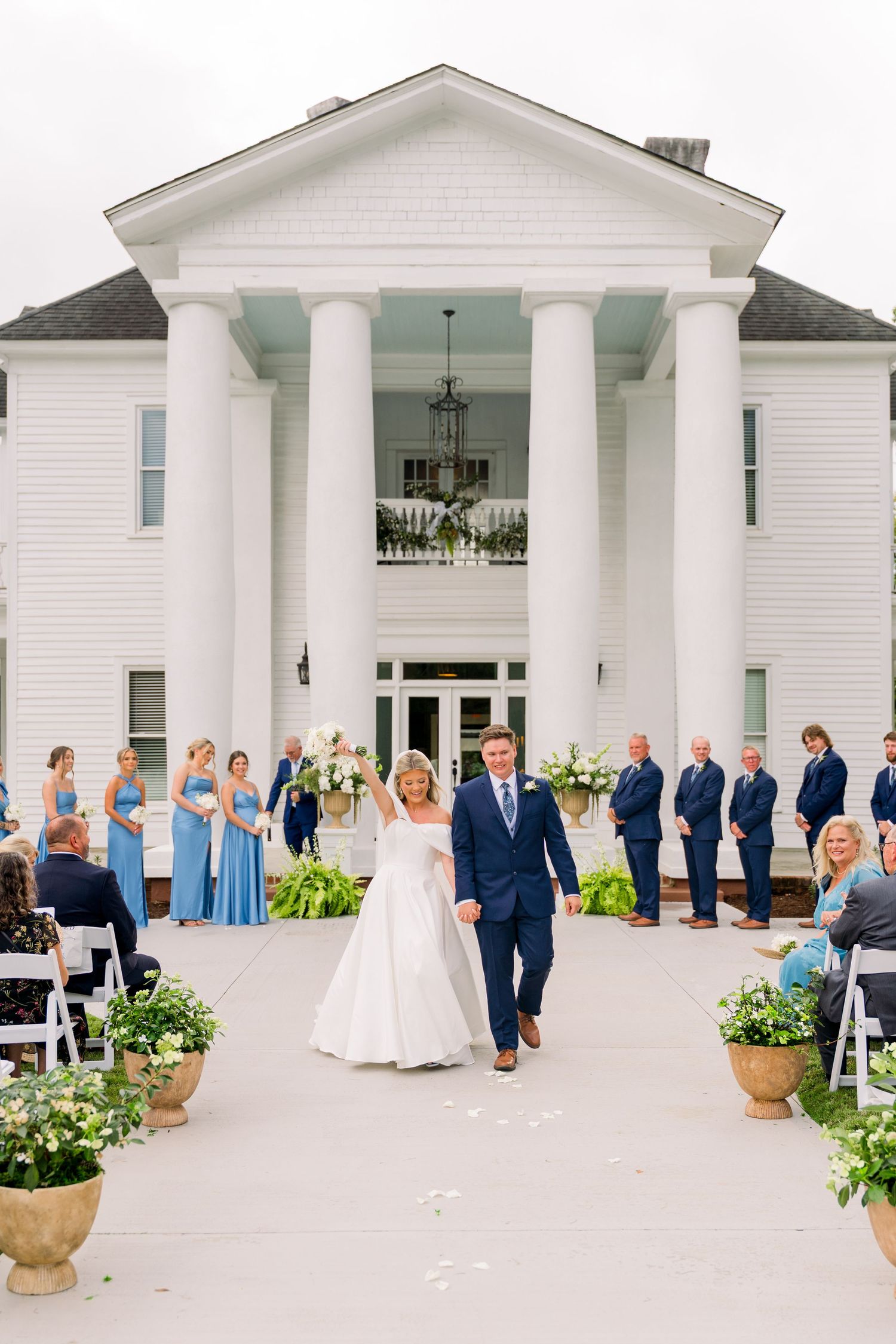 Bride and groom walk down the aisle at a white colonial wedding venue with blue bridesmaids and groomsmen.