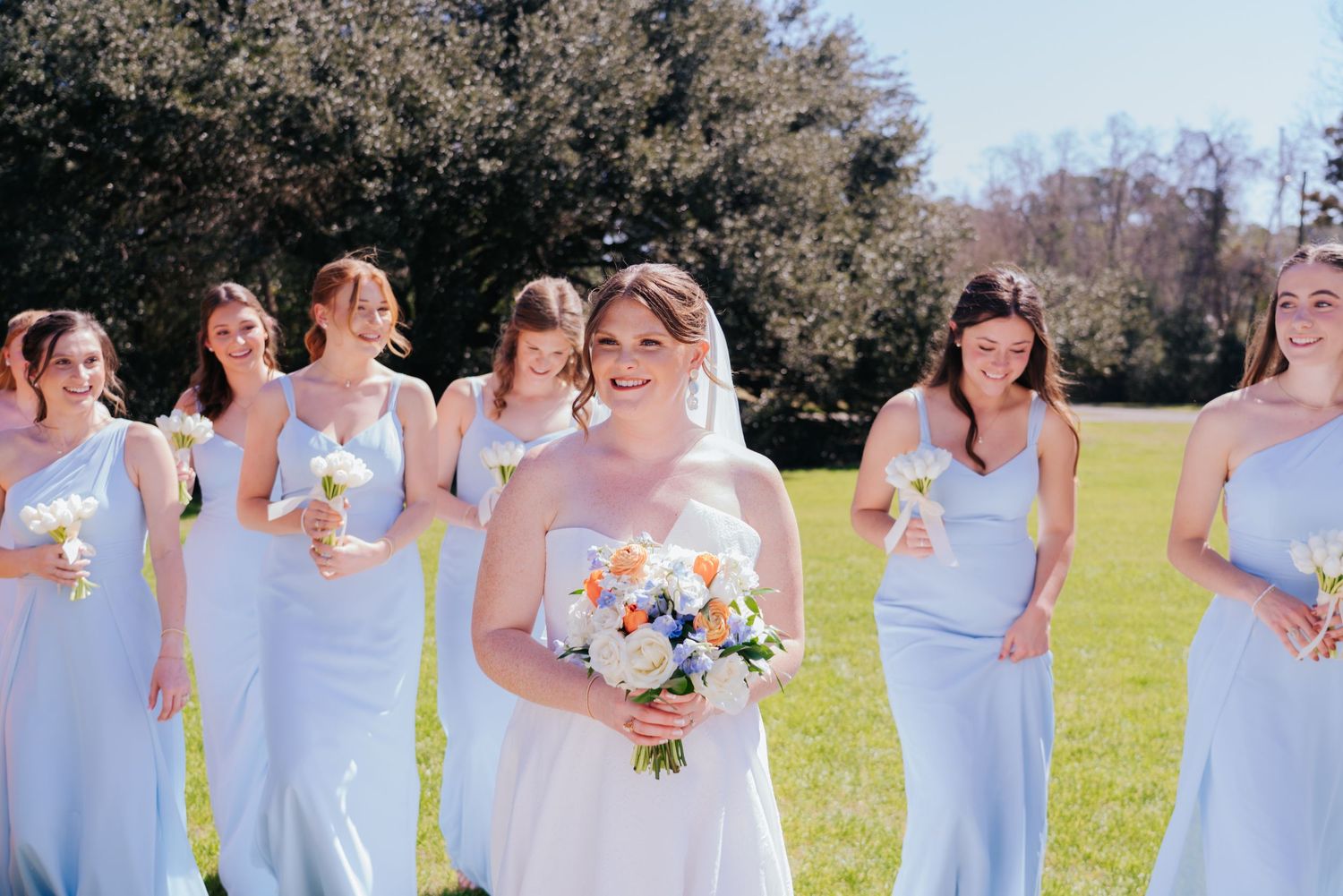 Bridesmaids in powder blue dresses gather outdoors in a sunny field.