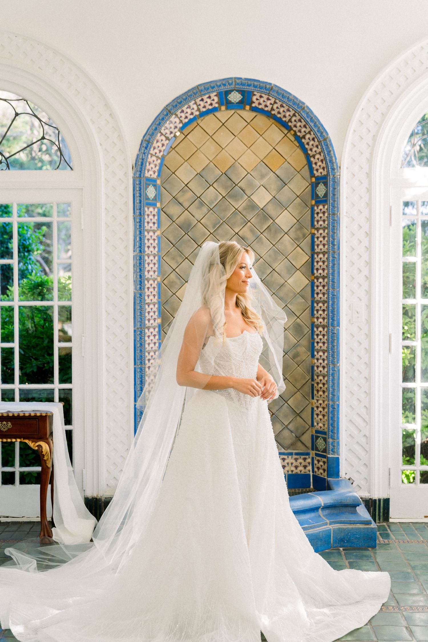 Bride in white gown and veil stands before ornate blue tiled arch in elegant sunlit room.