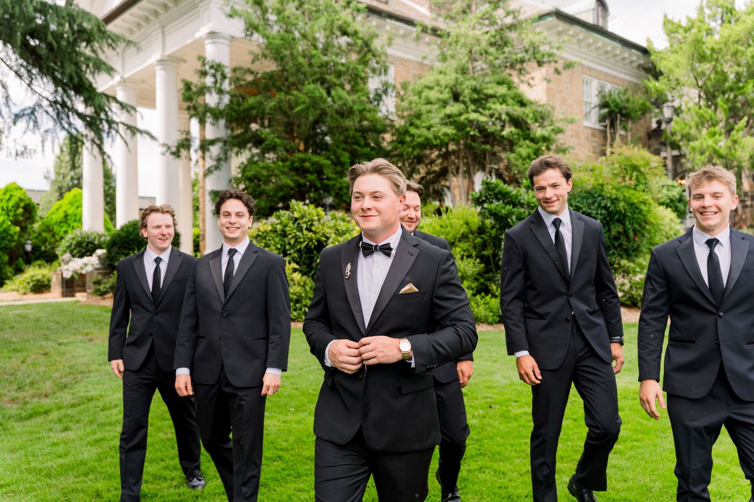 Groom in black tuxedo walks with smiling groomsmen in black suits on green lawn before elegant white mansion.