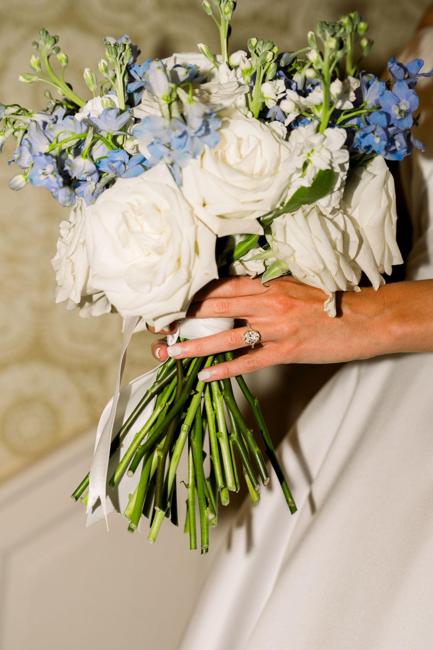 Bride holding a wedding bouquet of white roses and blue flowers with a diamond ring on her finger.