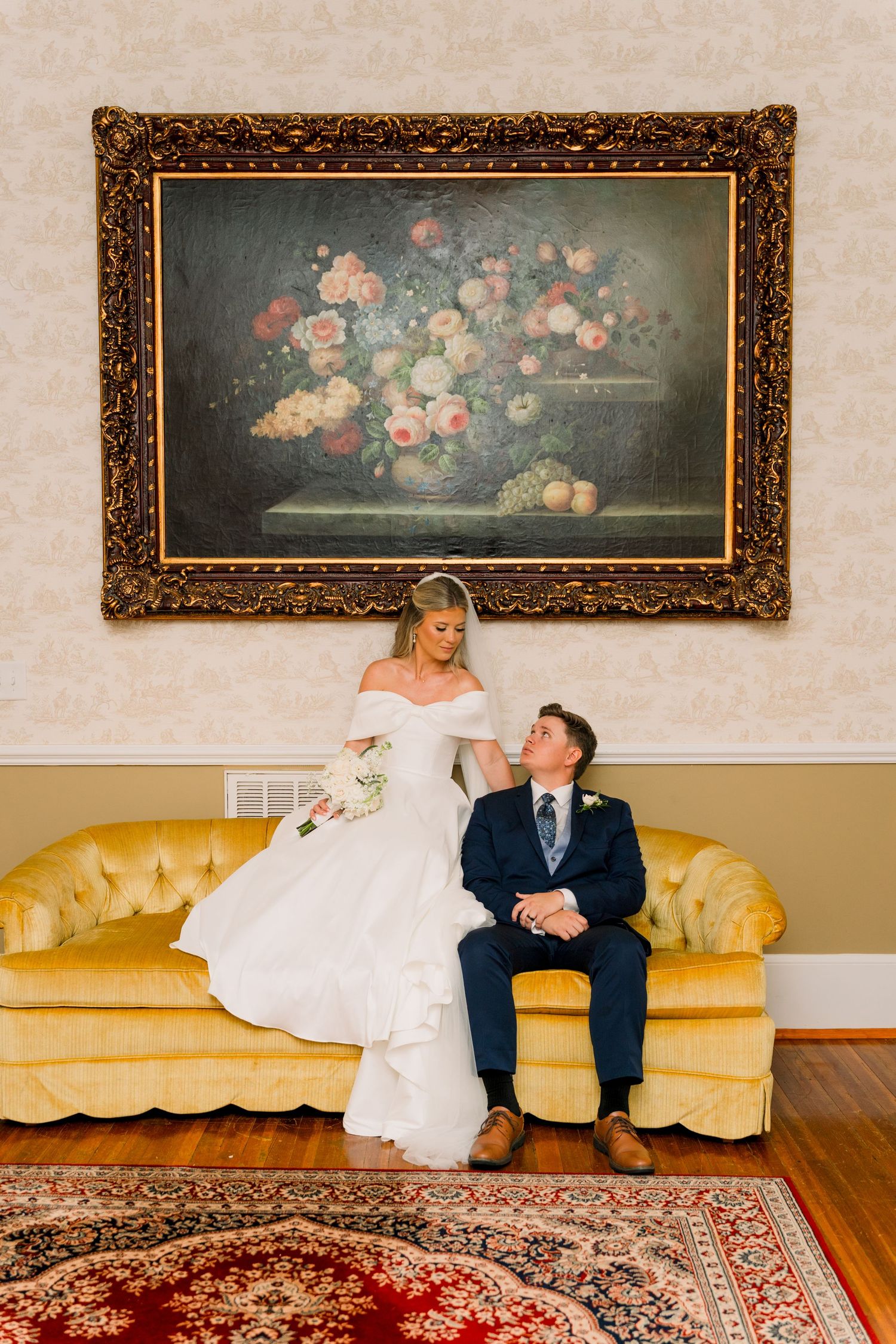 Bride and groom sitting on a vintage gold tufted sofa beneath an ornate floral oil painting in an elegant room.