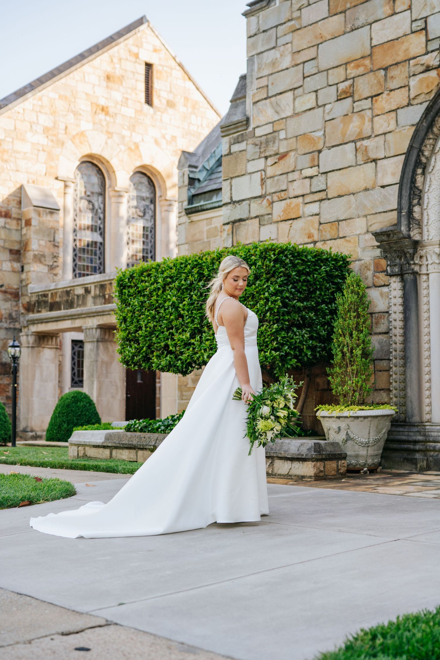 Bride in elegant white gown poses in front of stone church architecture with manicured gardens.