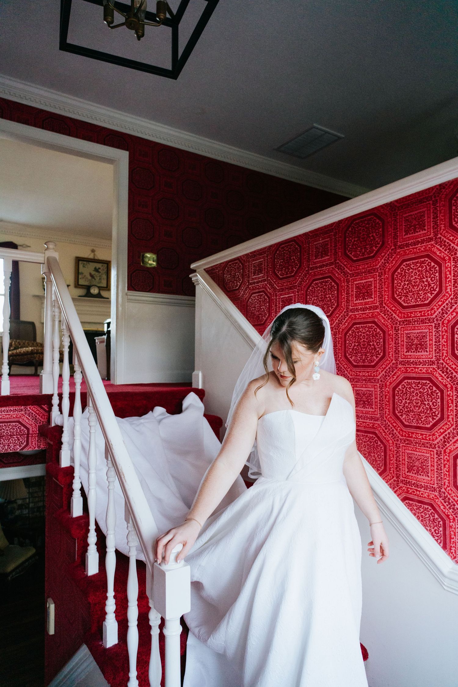 A bride descends a staircase with red wallpaper and white railings in her flowing gown.