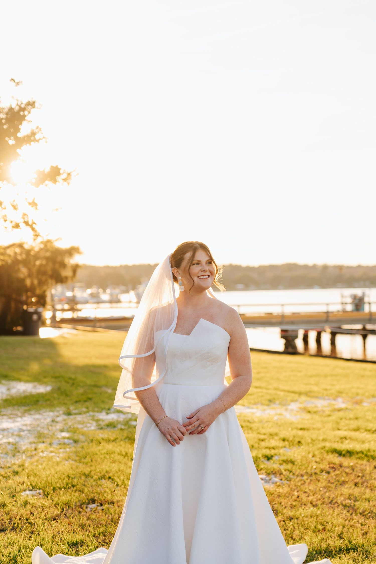 A wedding dress glows in golden sunlight near a waterfront dock.