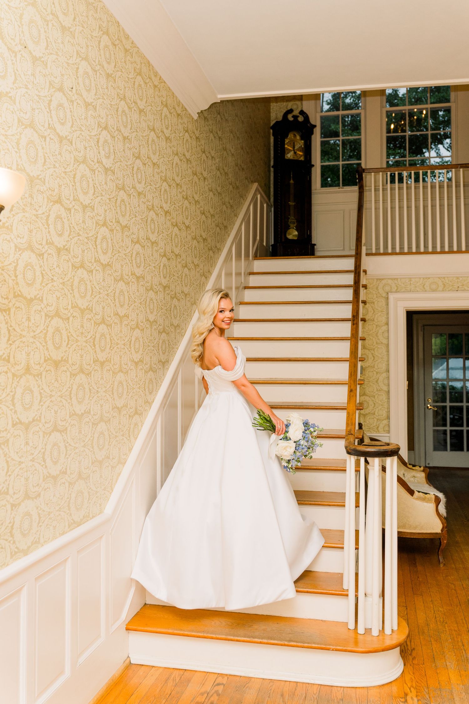 Bride in white off-shoulder gown holding bouquet on staircase with grandfather clock and patterned wallpaper.