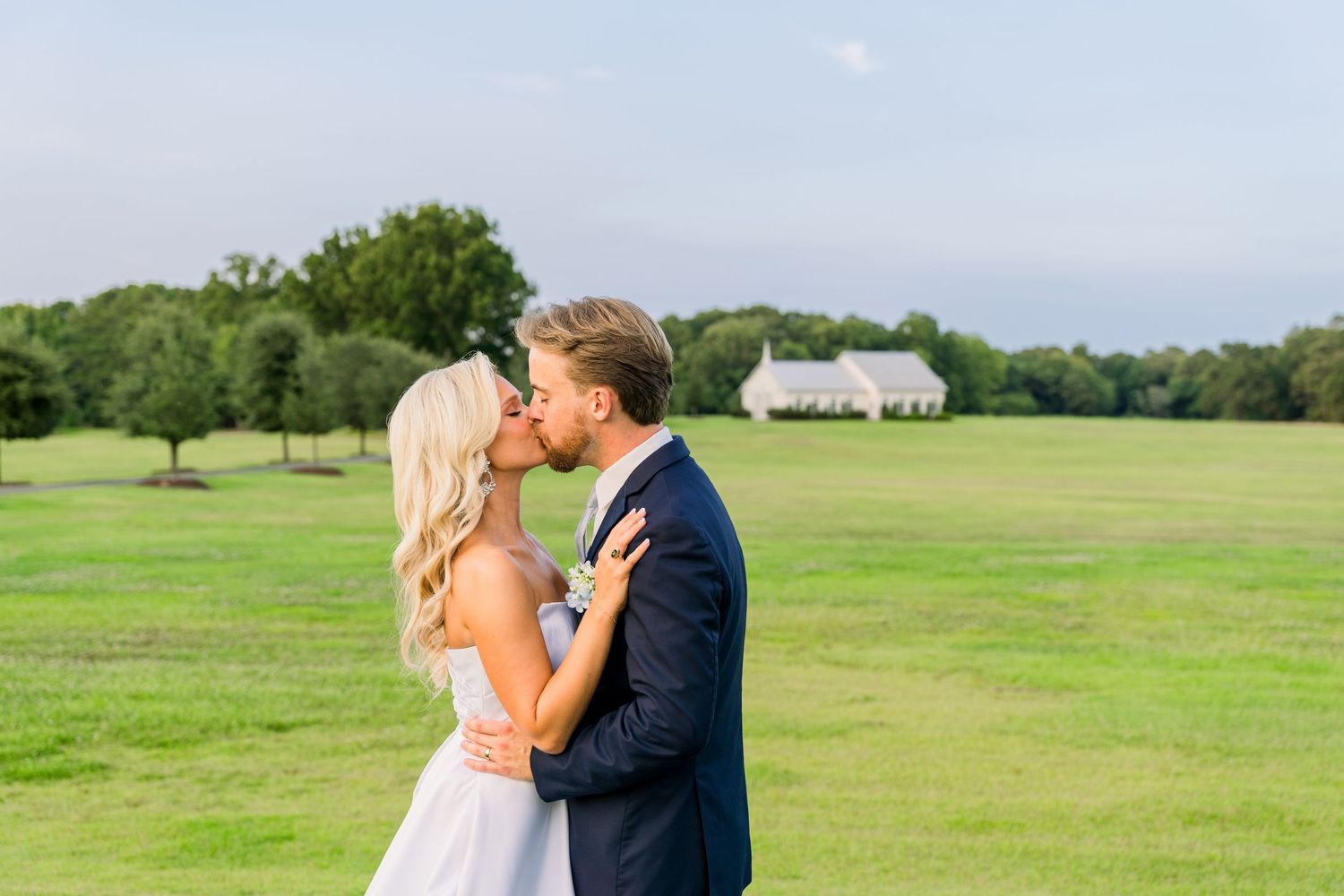 A bride and groom share a romantic kiss on a lush green lawn with a white estate in the background.