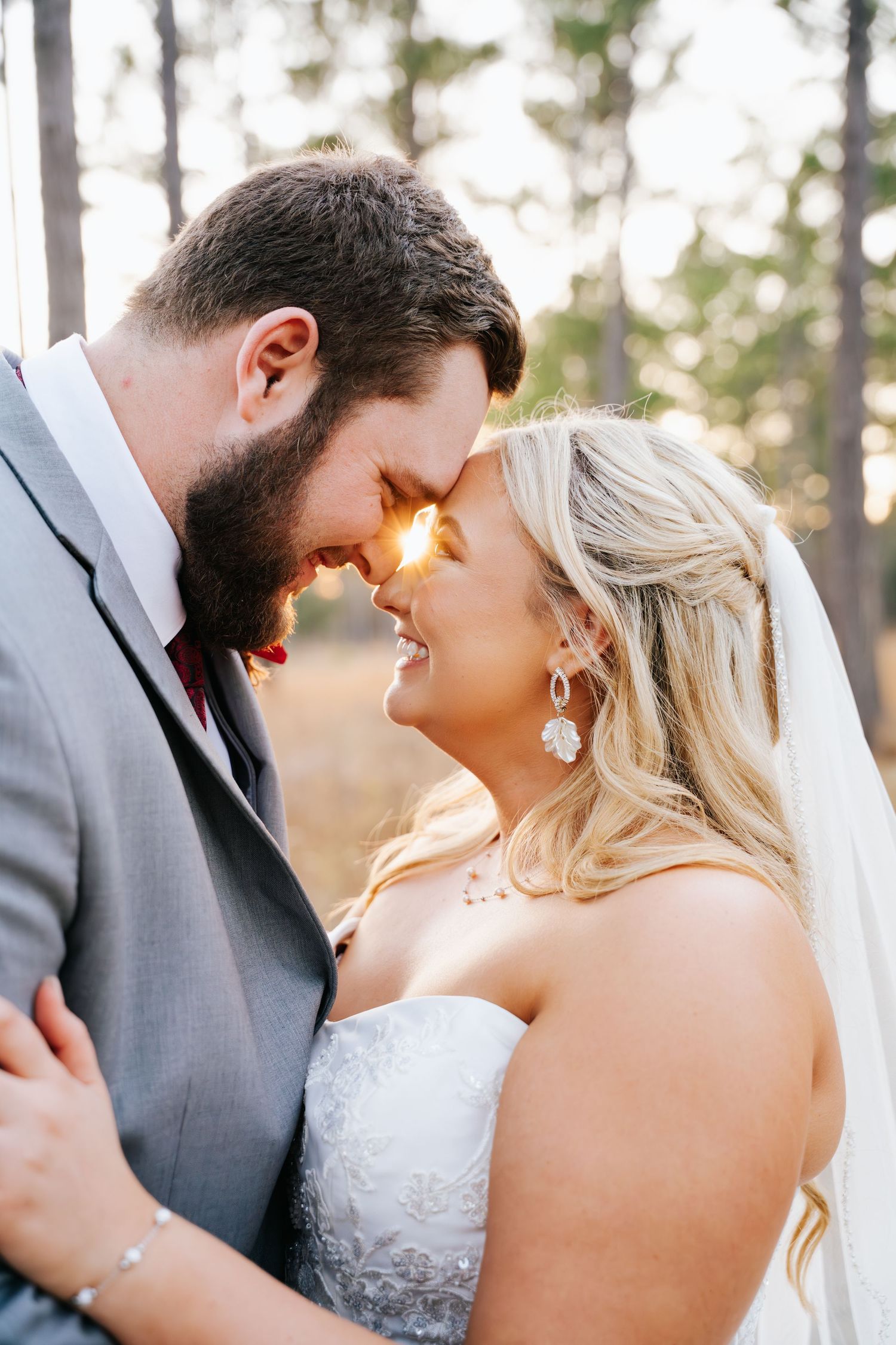 A bride and groom share an intimate forehead touch, smiling in golden sunlight, with the bride wearing a veil and floral earrings.