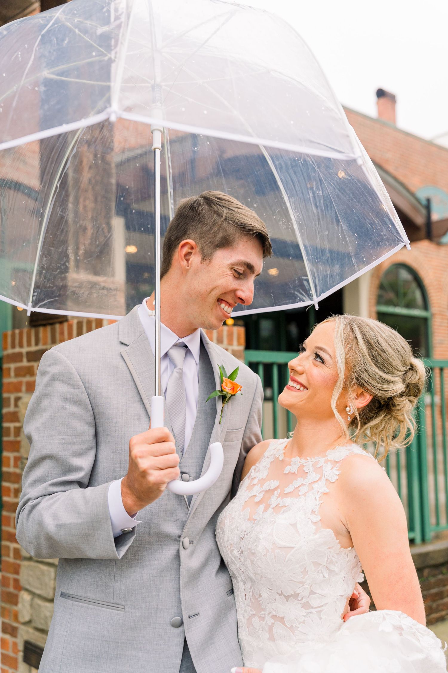 Bride and groom smiling at each other under a clear umbrella on their rainy wedding day.