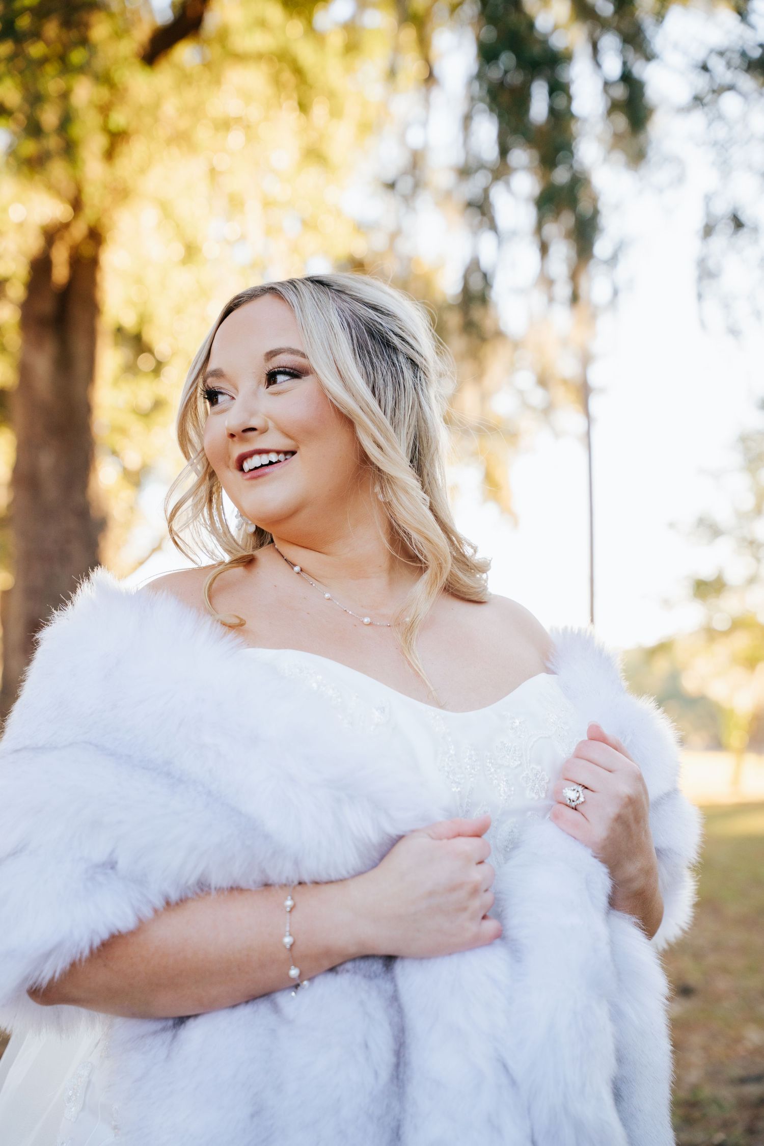 A bride wrapped in a white fur shawl smiles while looking up on a sunny autumn day in an outdoor setting.
