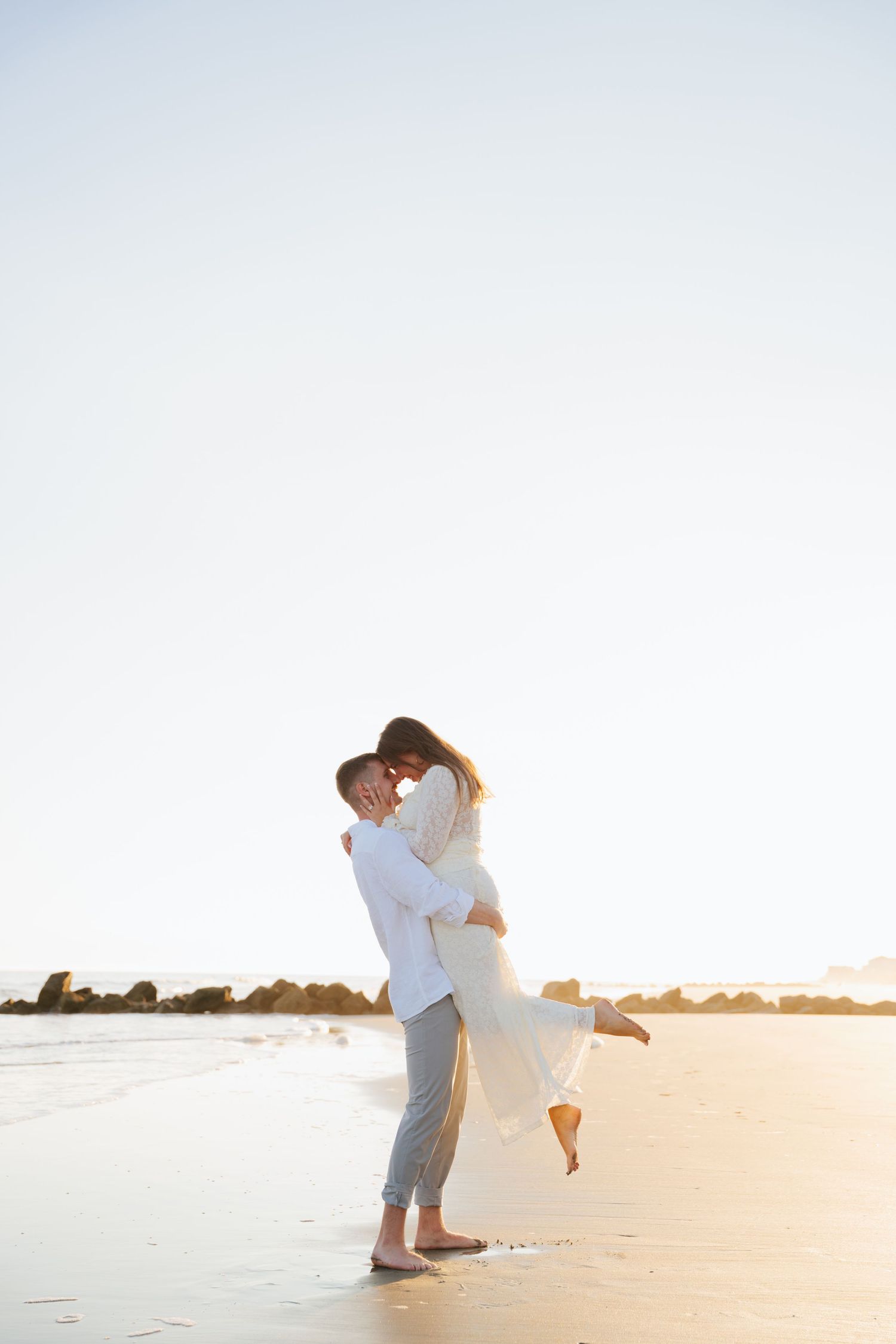 Romantic couple embraces on sandy beach at sunset with person lifted in white dress and grey pants.