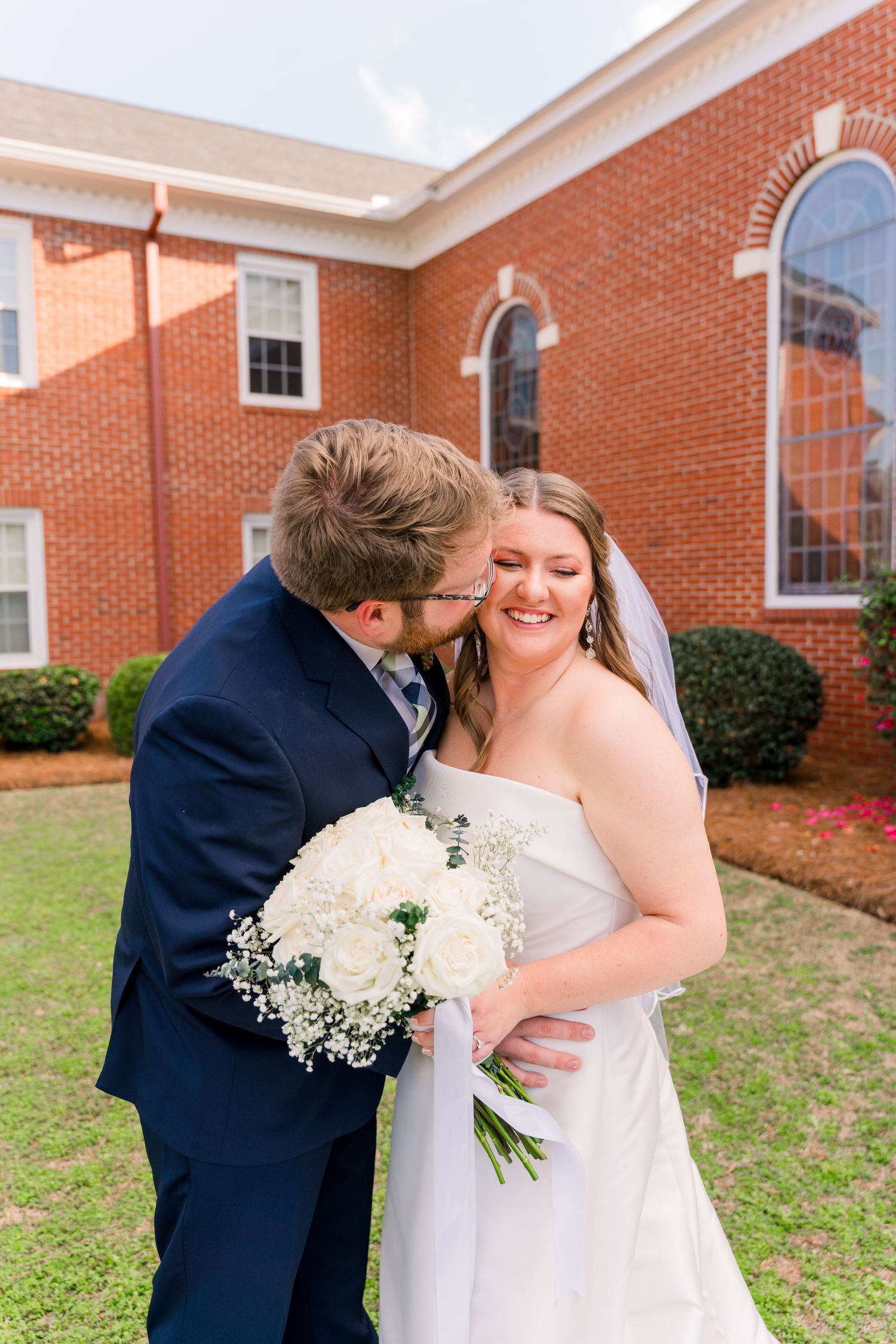 Groom in navy suit kisses bride's cheek as she smiles holding white rose bouquet outside brick church.