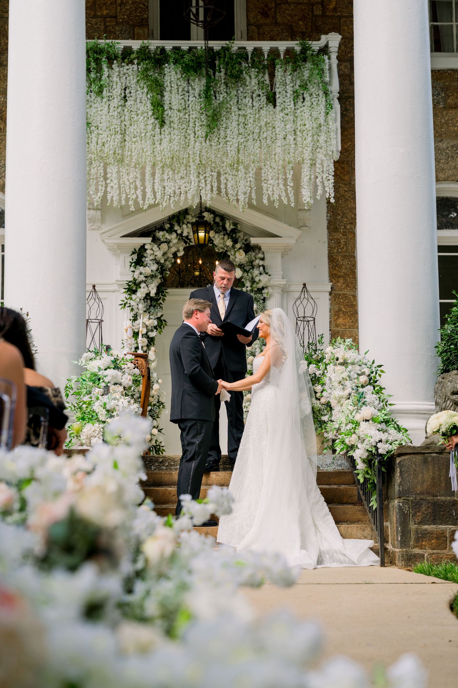 Bride and groom exchange vows outdoors at elegant venue with white floral arch and cascading greenery backdrop.