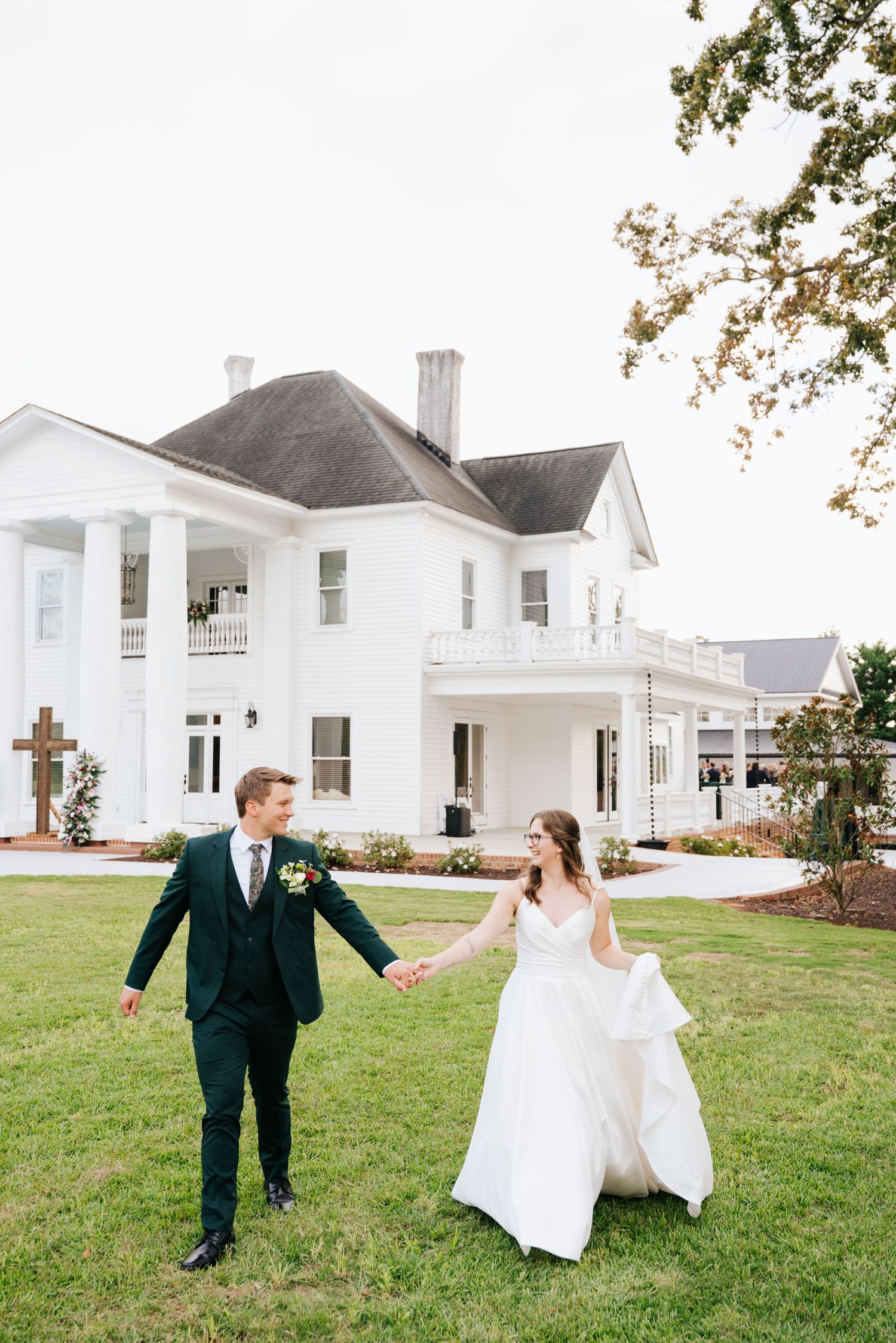 A bride and groom hold hands walking on green grass in front of a grand white Southern mansion on their wedding day.