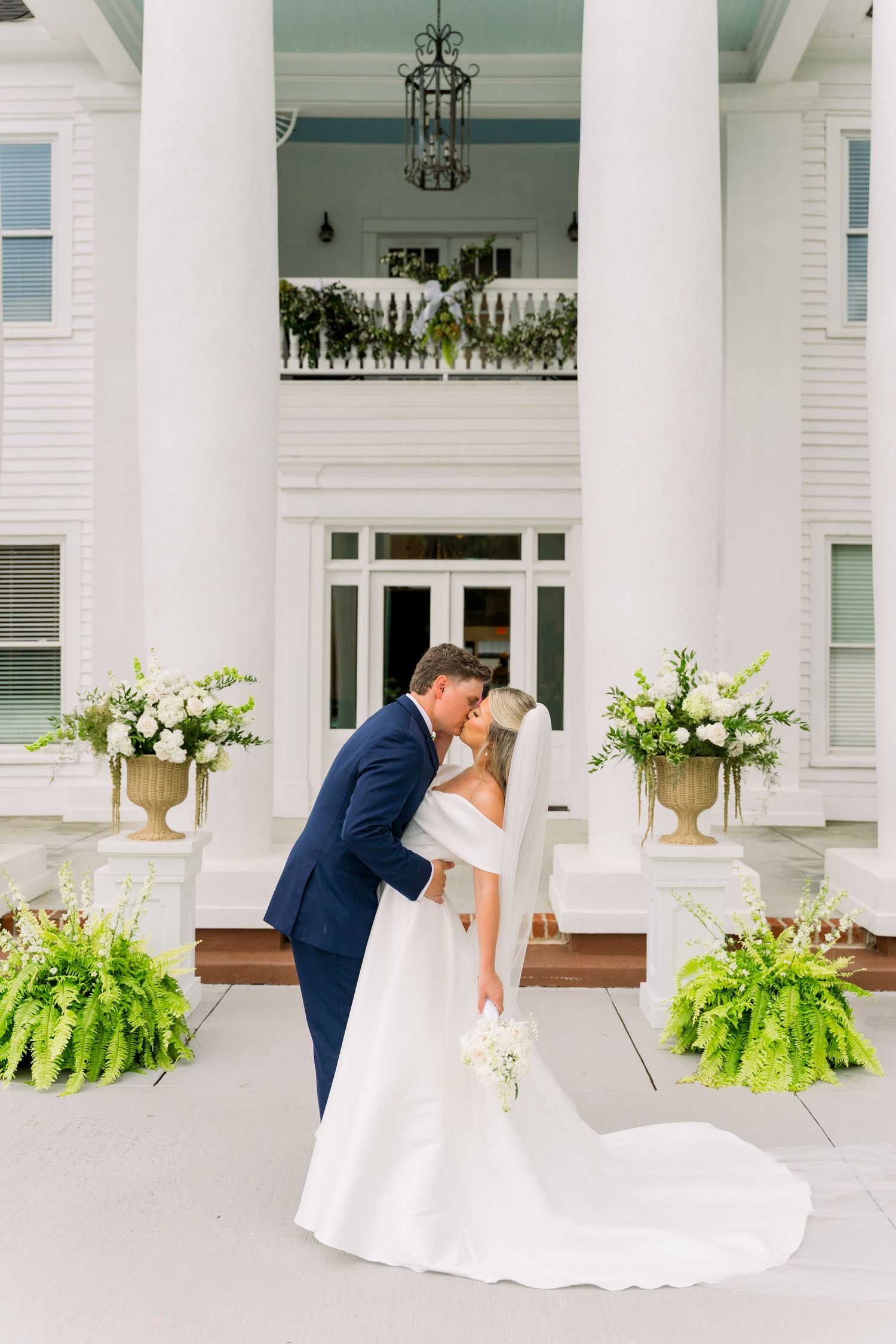 Bride and groom share a romantic kiss in front of an elegant white Southern mansion with floral arrangements.