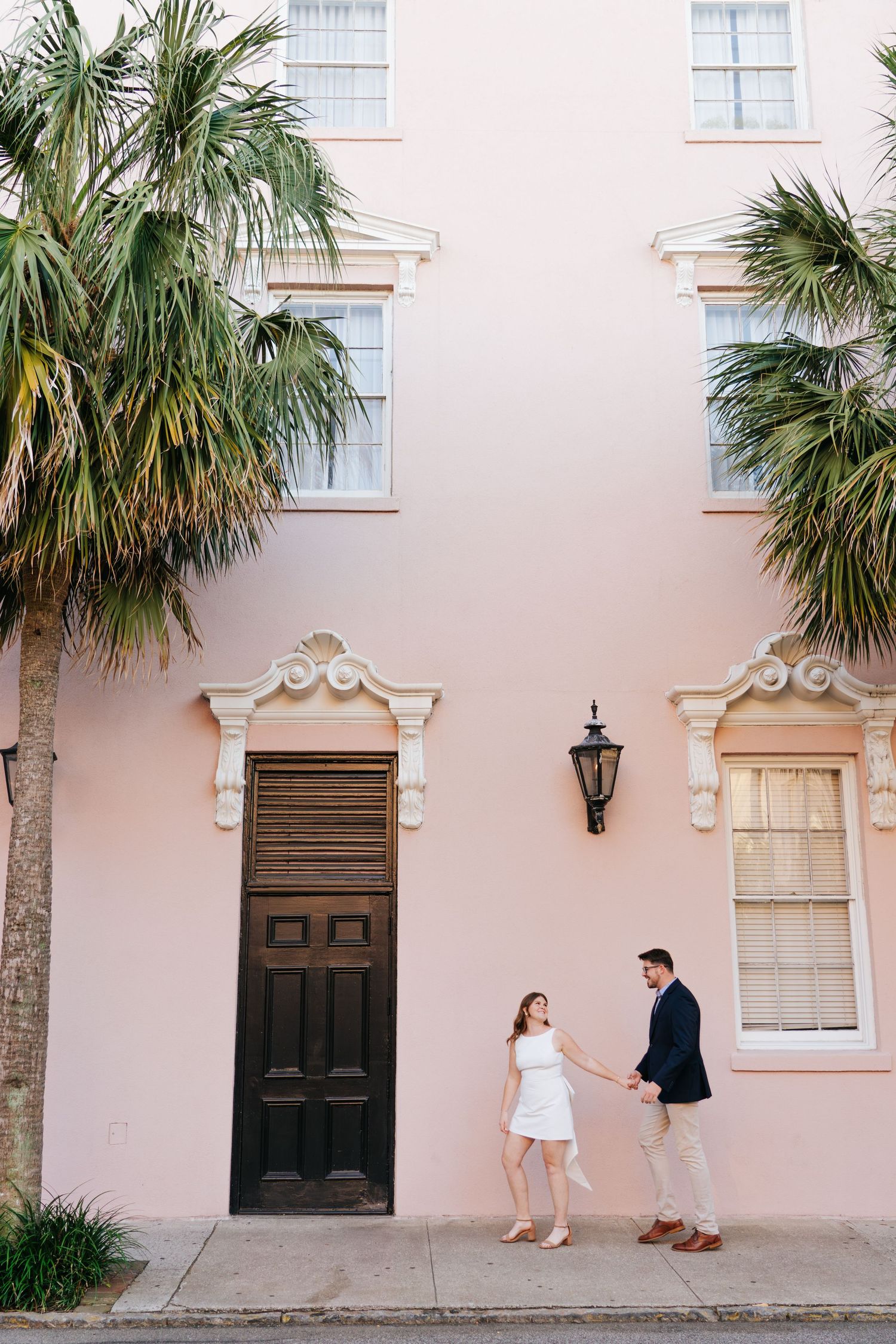 A couple stands in front of a pink Charleston-style building with palm trees and ornate architectural details.