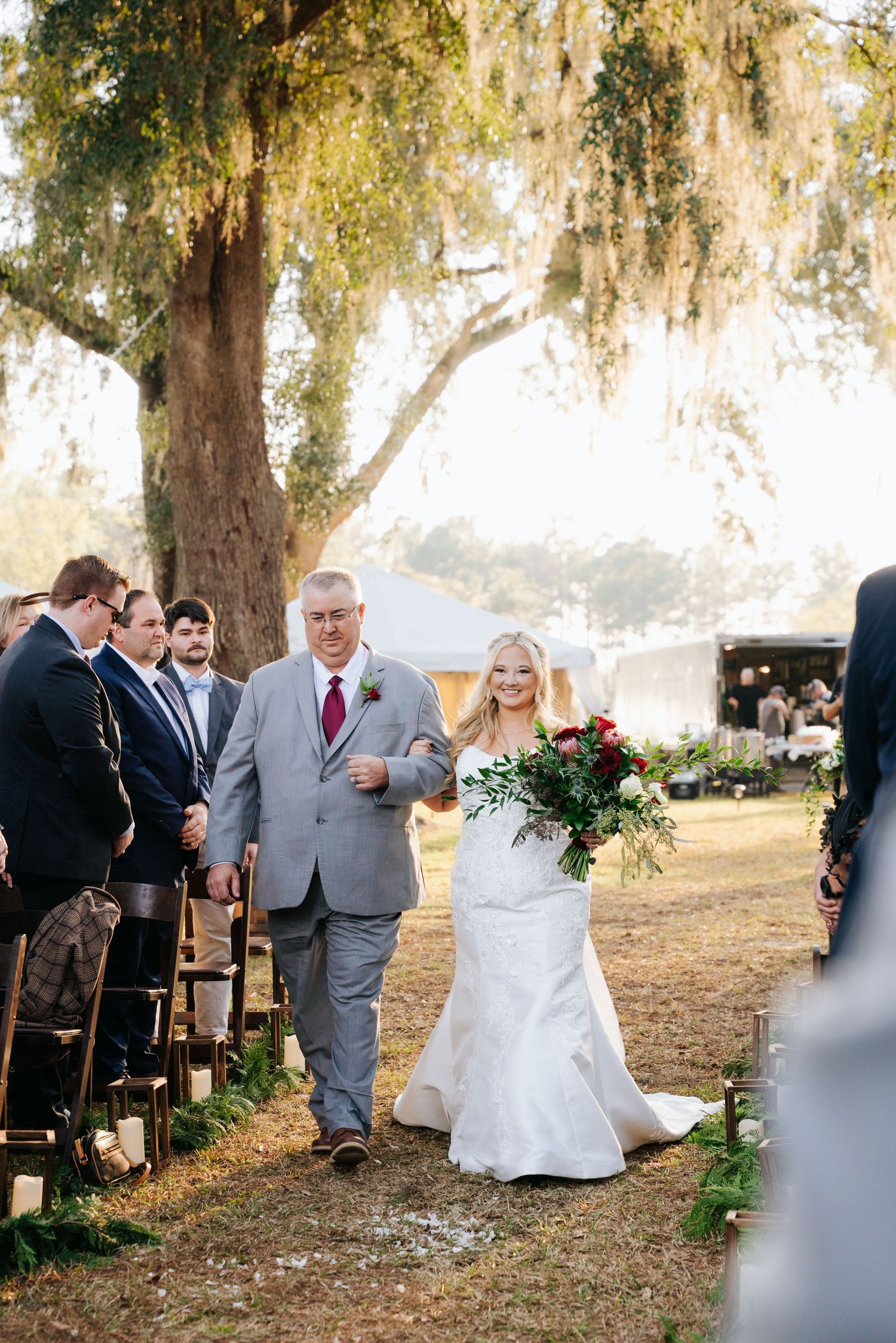 Outdoor wedding ceremony under moss-draped oak trees with guests seated on wooden chairs in a natural setting.