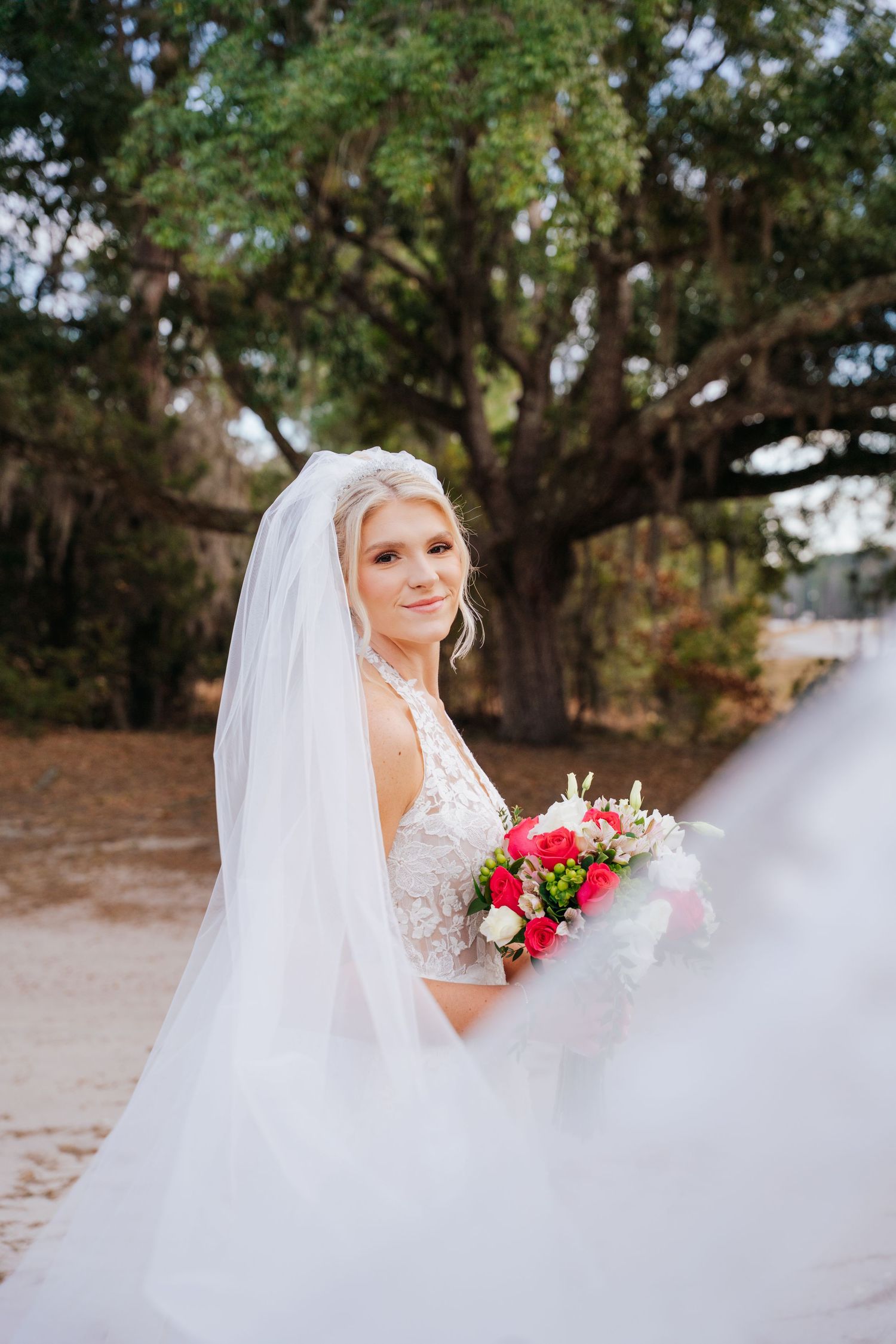 A bride in a white lace dress holds a vibrant red bouquet while posing outdoors under large oak trees.