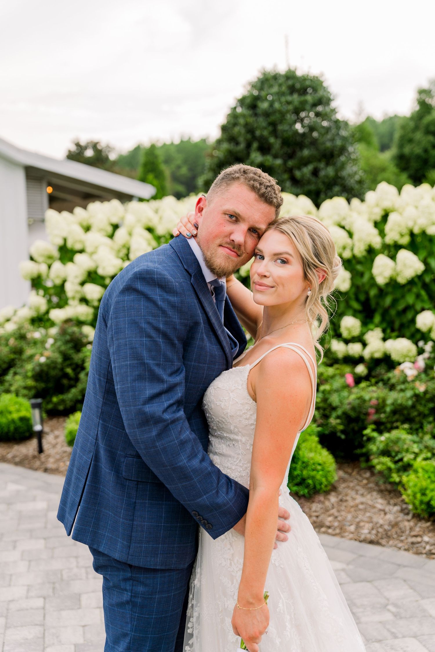 Bride and groom embracing in front of white hydrangeas, wearing a blue suit and lace wedding gown outdoors.