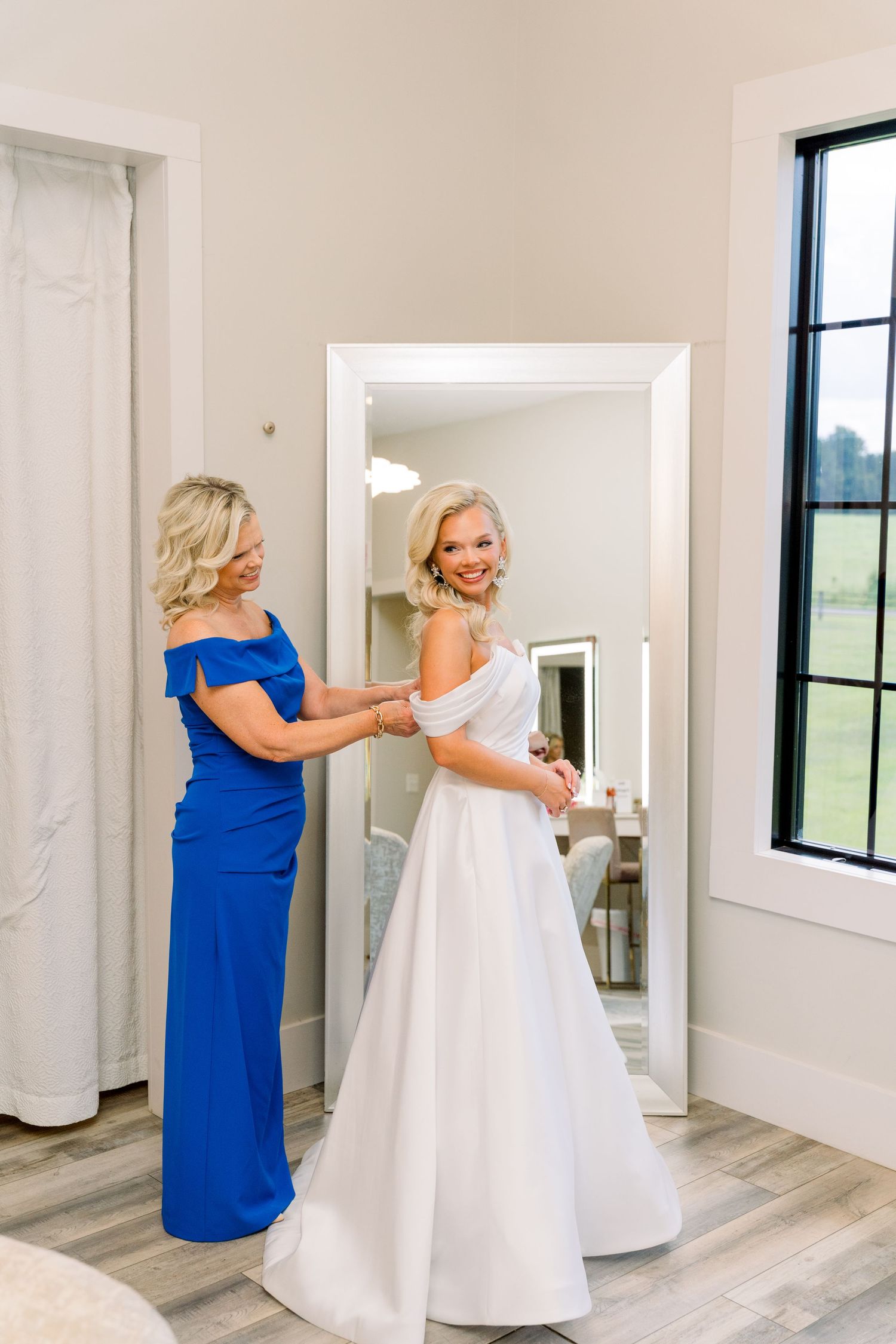 Bride in white off-shoulder gown gets help from woman in royal blue dress before wedding ceremony.