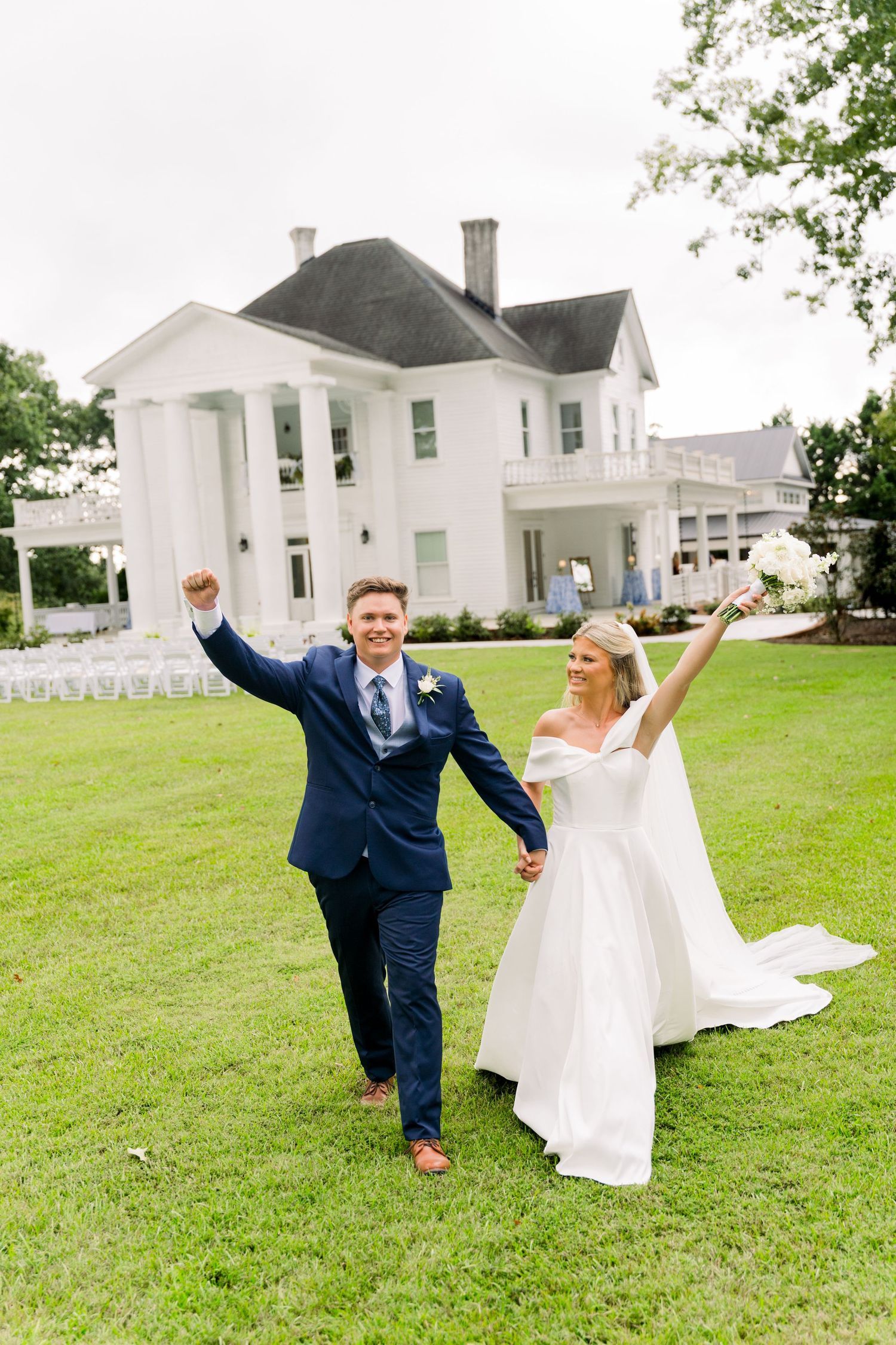 Joyful newlyweds celebrate on green lawn before white colonial mansion, bride in gown holding bouquet, groom in navy suit.