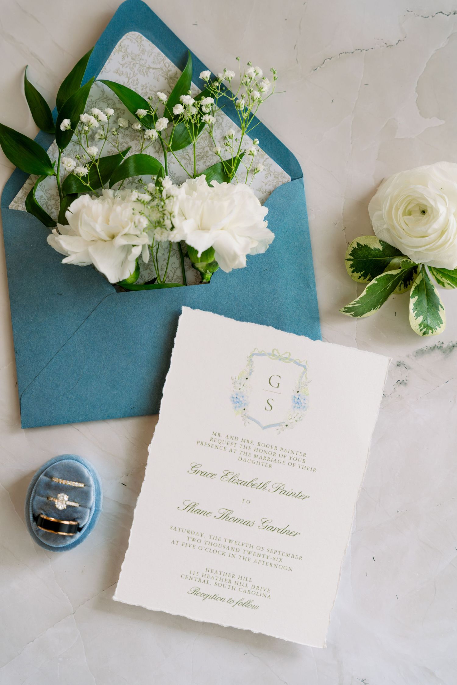 Elegant wedding invitation suite with teal envelope, white peonies, ranunculus, and a diamond ring box on marble.