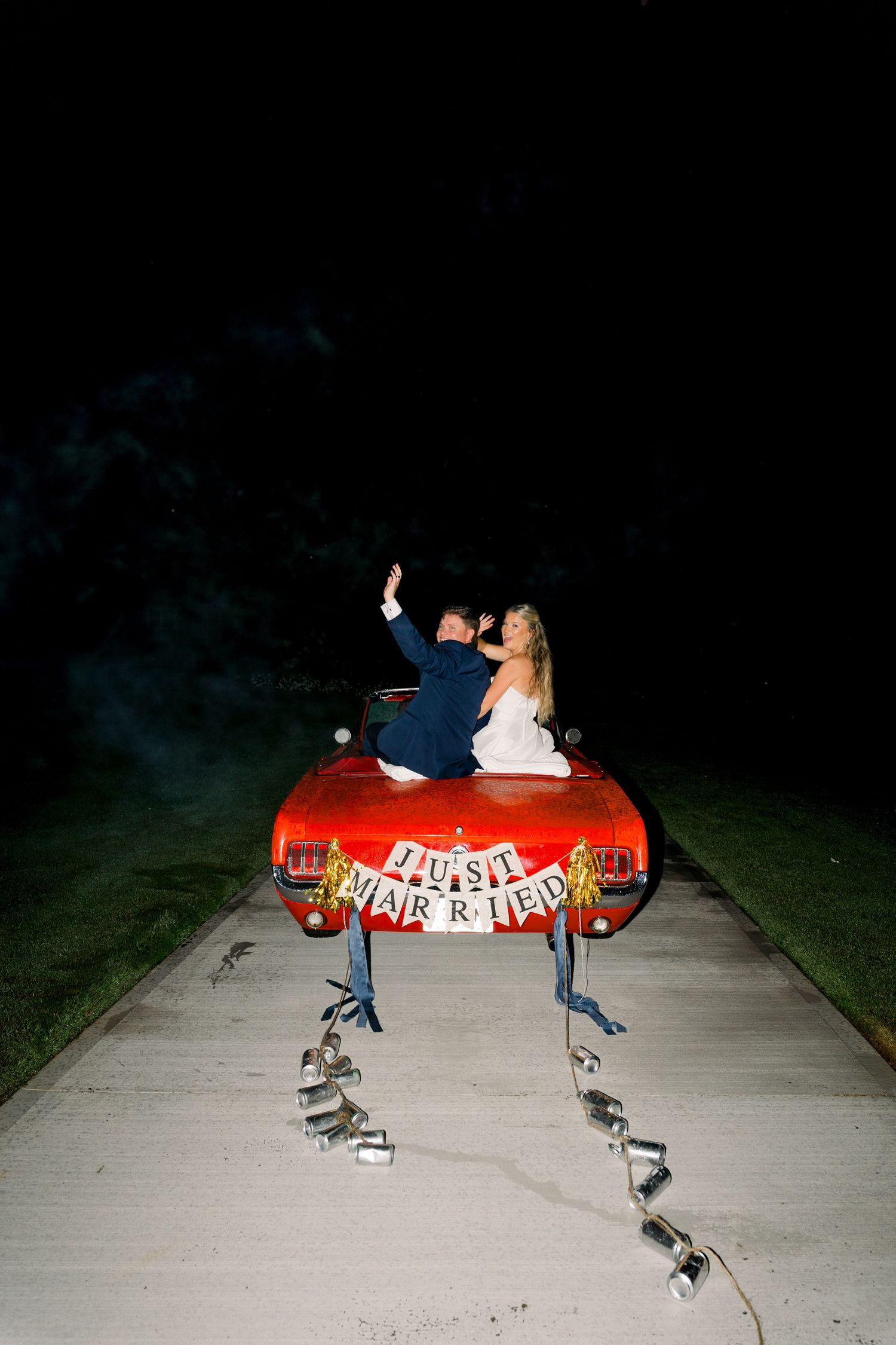 Newlyweds ride in a red convertible with Just Married banner and tin cans trailing at night.
