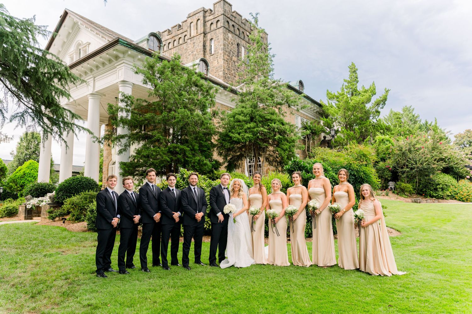 Wedding party poses in front of elegant stone mansion, bridesmaids in champagne gowns with groomsmen in black suits.