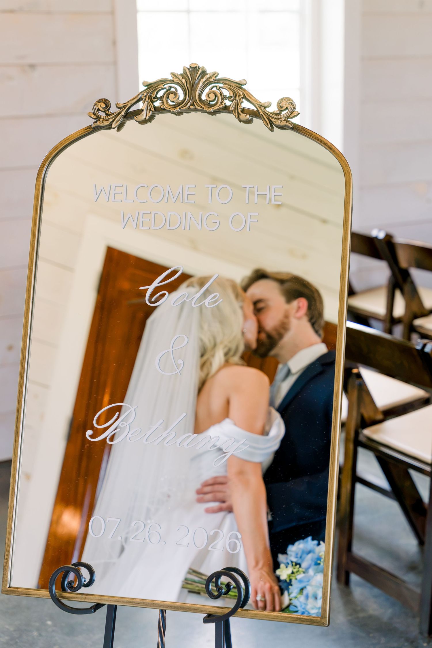 Gold ornate mirror wedding welcome sign displaying Cole and Brittany's wedding date 07.26.2026 with couple kissing in reflection.