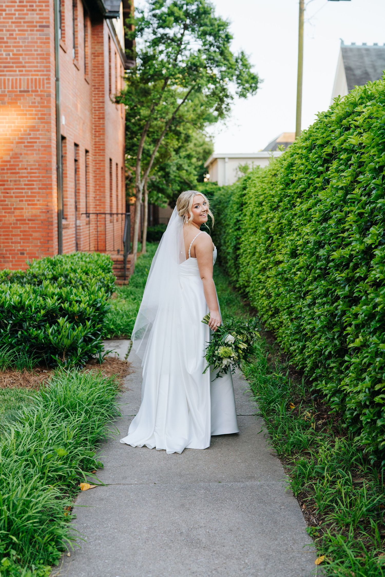 A bride in a white dress stands on a pathway between brick buildings and green hedges.