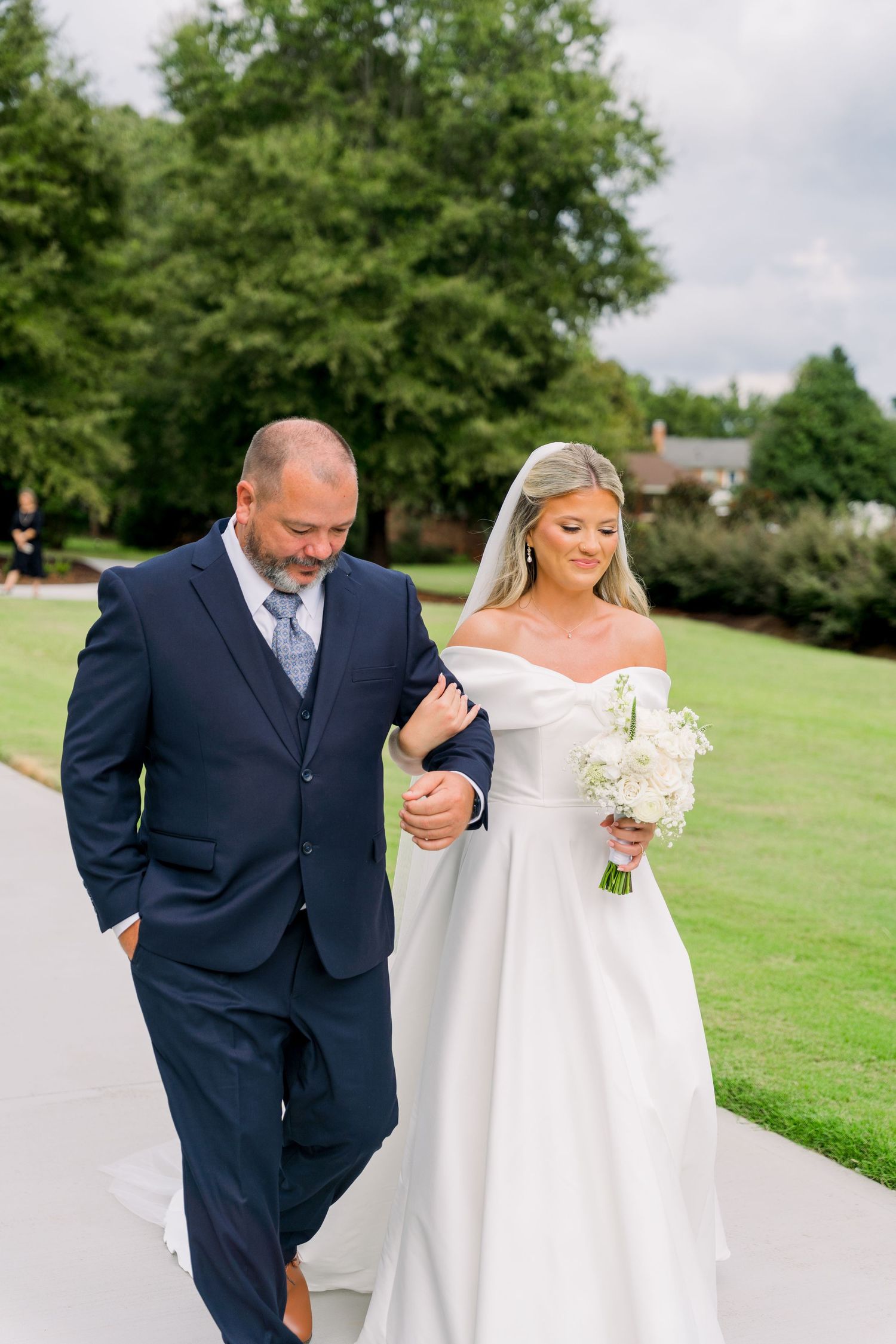 Bride in off-shoulder gown holding white bouquet walks arm-in-arm with man in navy suit on outdoor path.