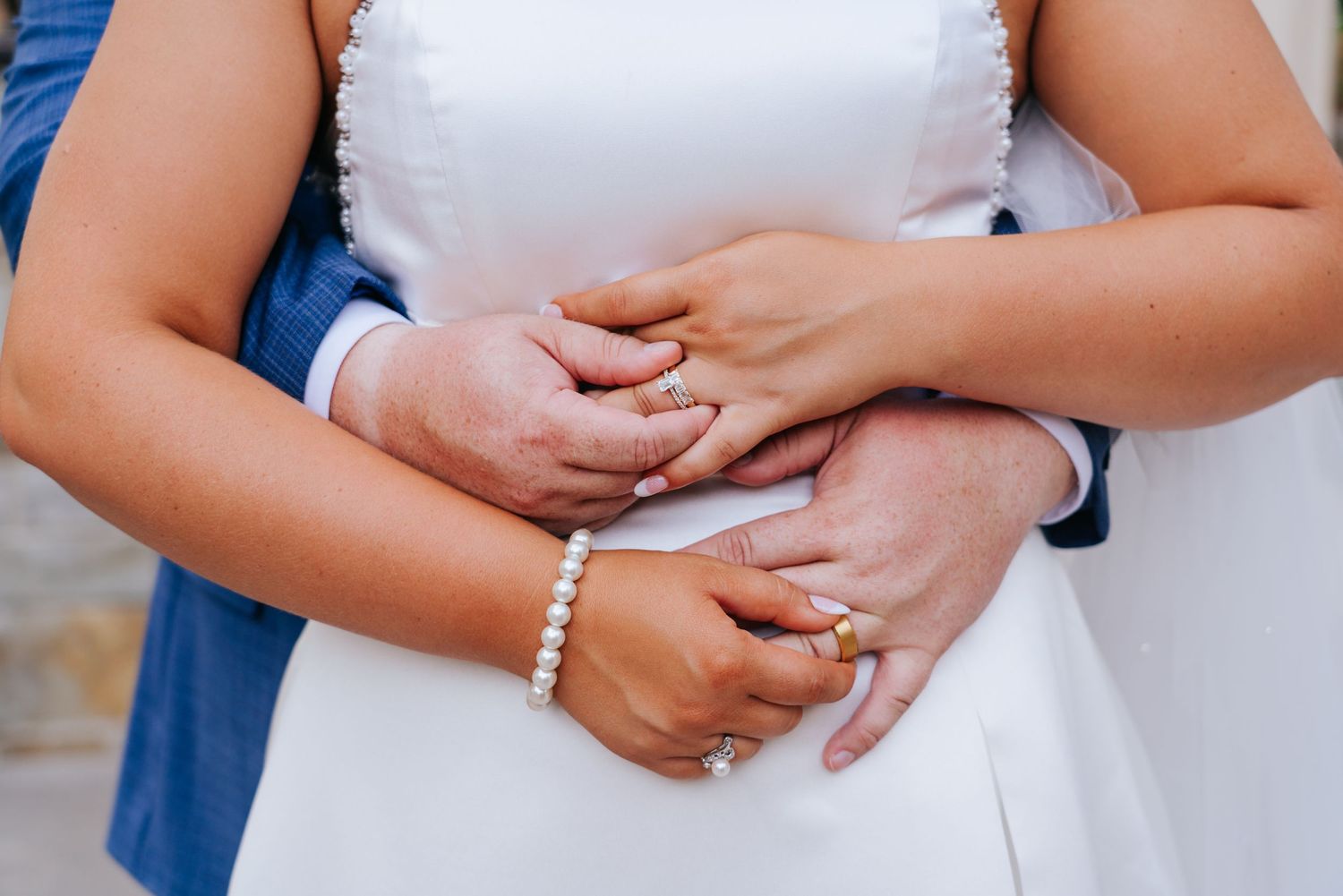 Close-up detail of hands with wedding rings and bridal jewelry against white dress.