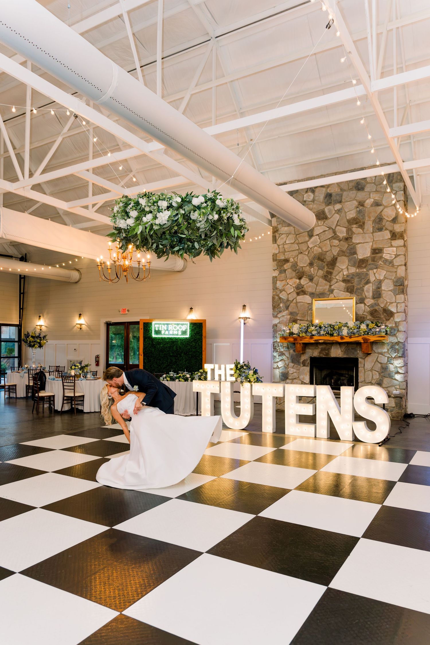 Newlyweds share a romantic dip kiss on a black and white checkered dance floor beside large marquee letters spelling The Tutens.
