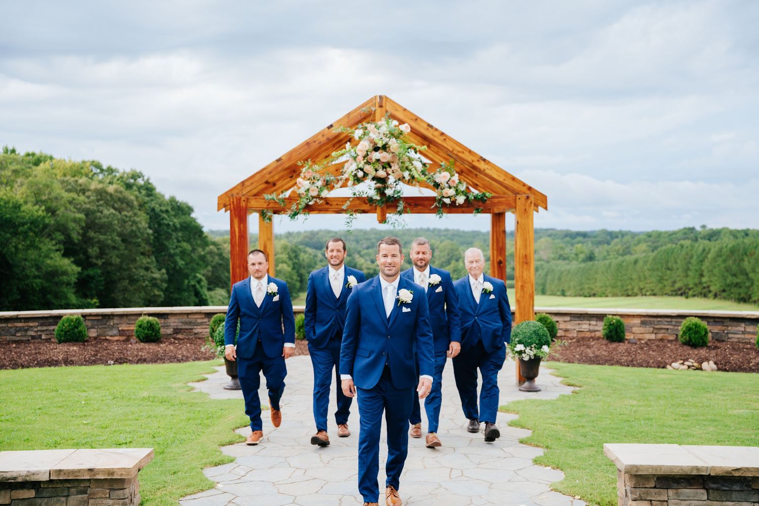 Groomsmen in navy blue suits walk together under a wooden pavilion at an outdoor wedding venue.