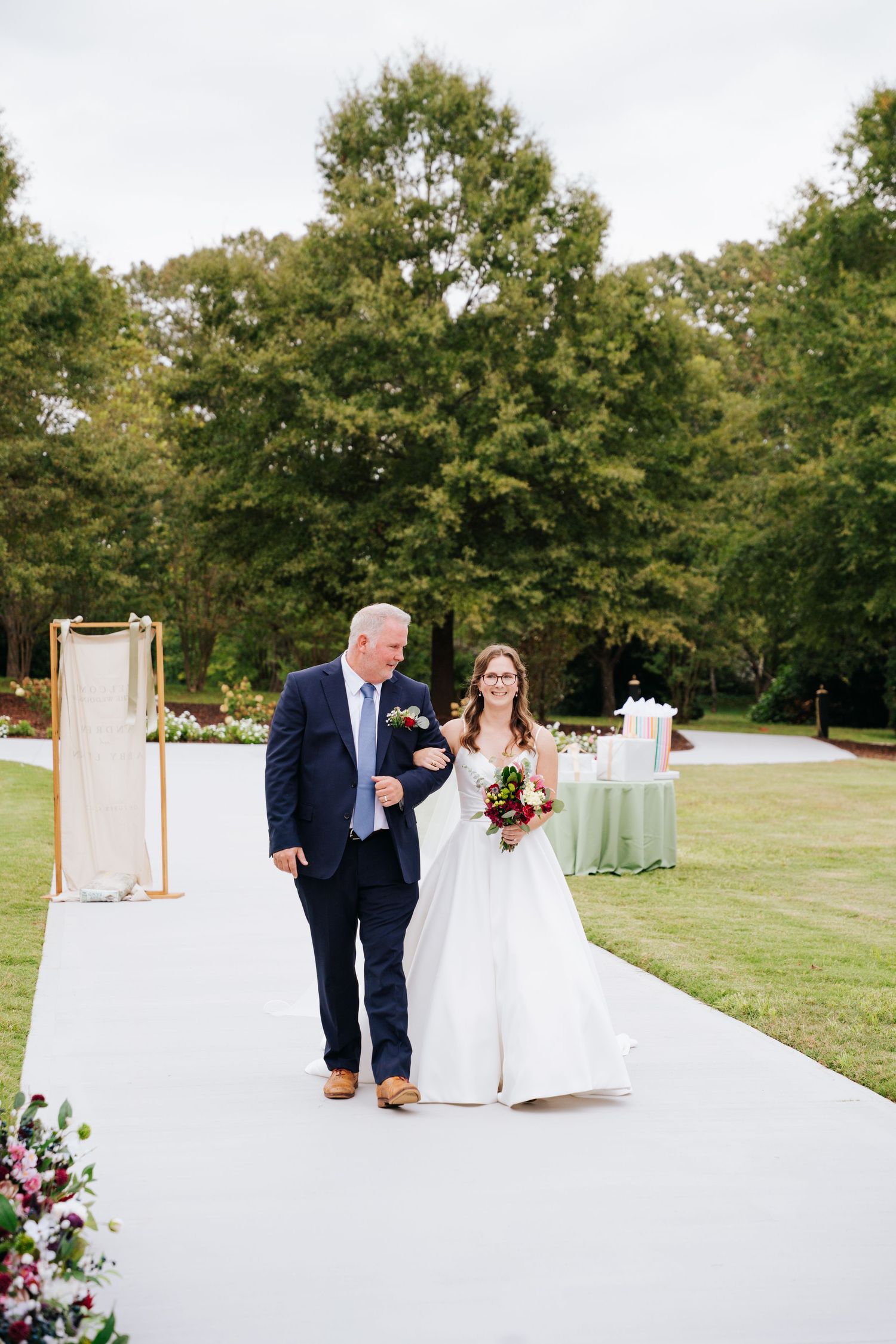 A white aisle runner stretches across a green lawn with trees in the background at an outdoor wedding ceremony.