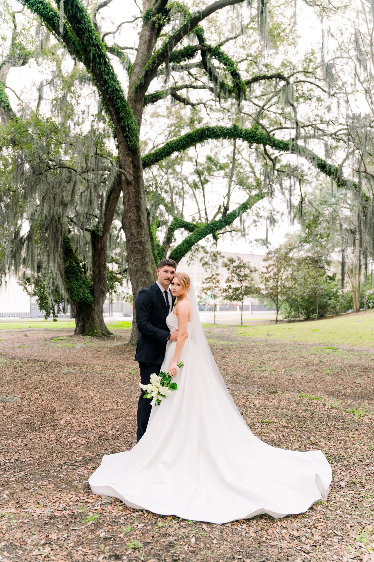 A romantic wedding couple poses under moss-draped oak trees in a Southern outdoor setting.