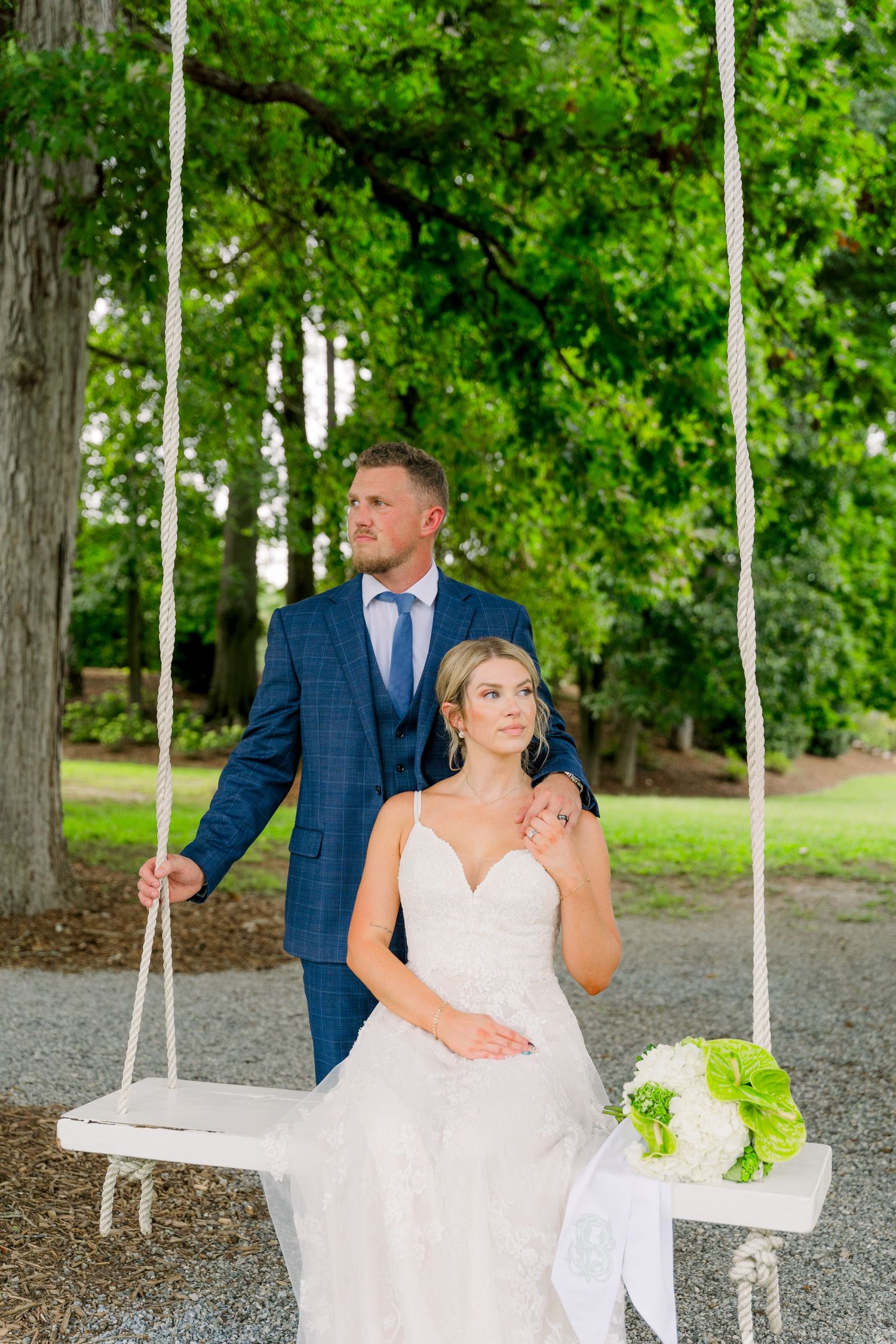 Bride sitting on a white rope swing holding a bouquet while groom in blue suit stands behind her outdoors.
