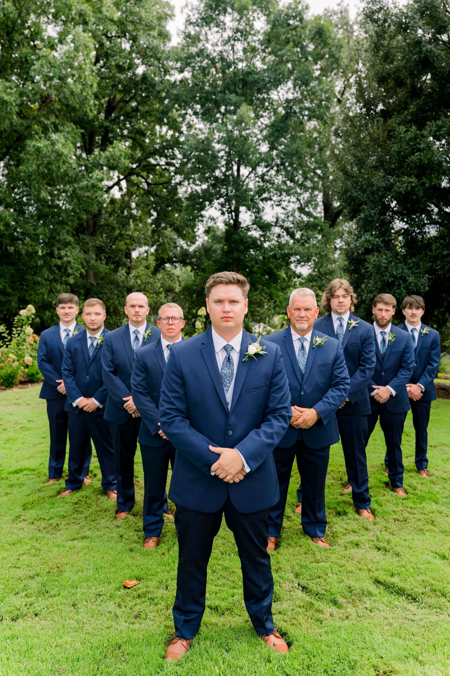Groom in navy blue suit stands in front of groomsmen in matching suits outdoors in a lush garden setting.