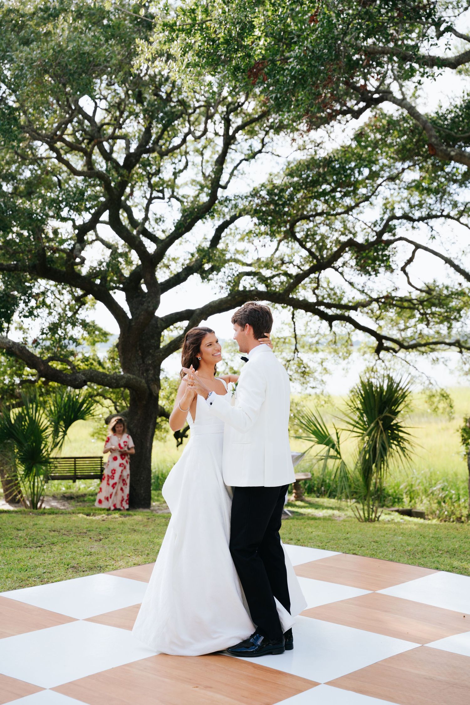 Bride and groom share first dance on checkered outdoor dance floor beneath sprawling oak tree at elegant garden wedding.