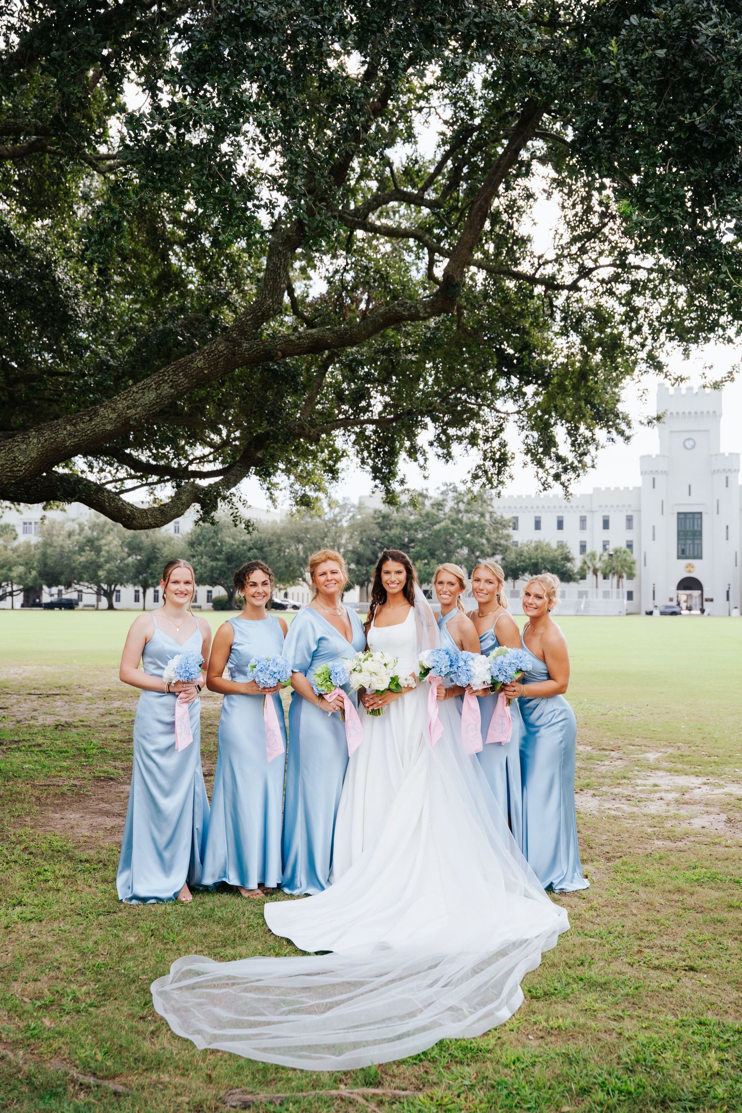 Wedding party in light blue dresses poses under oak trees on sunny grounds.