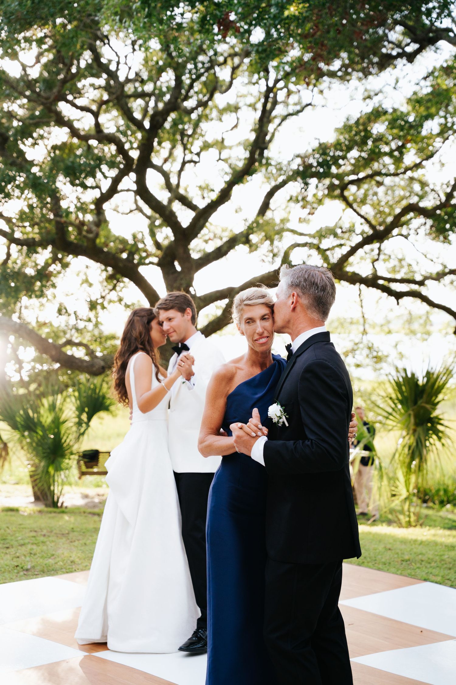 Bride and groom dancing together with bride's mom and dad on a checkered dance floor beneath oak trees.