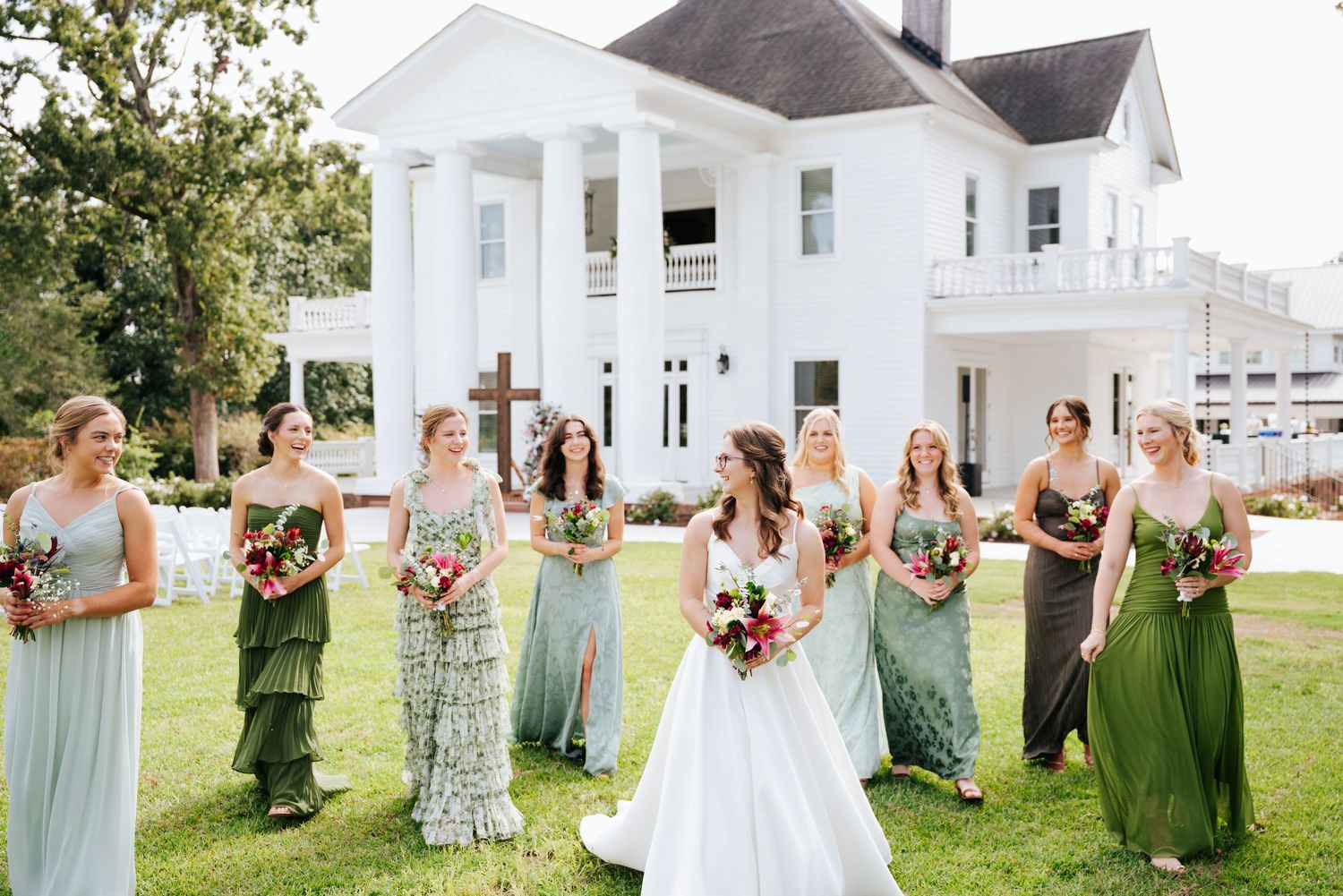 Wedding party in green dresses poses together on a sunny lawn in front of a white building.