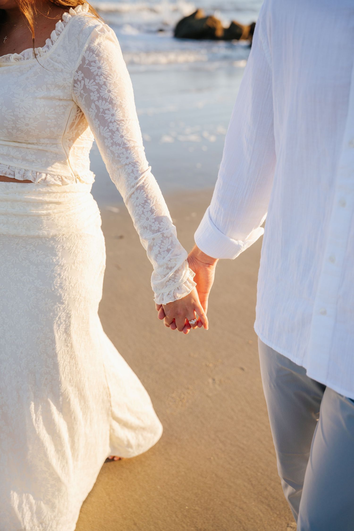 Two people in white outfits hold hands while walking on a sandy beach at sunset.