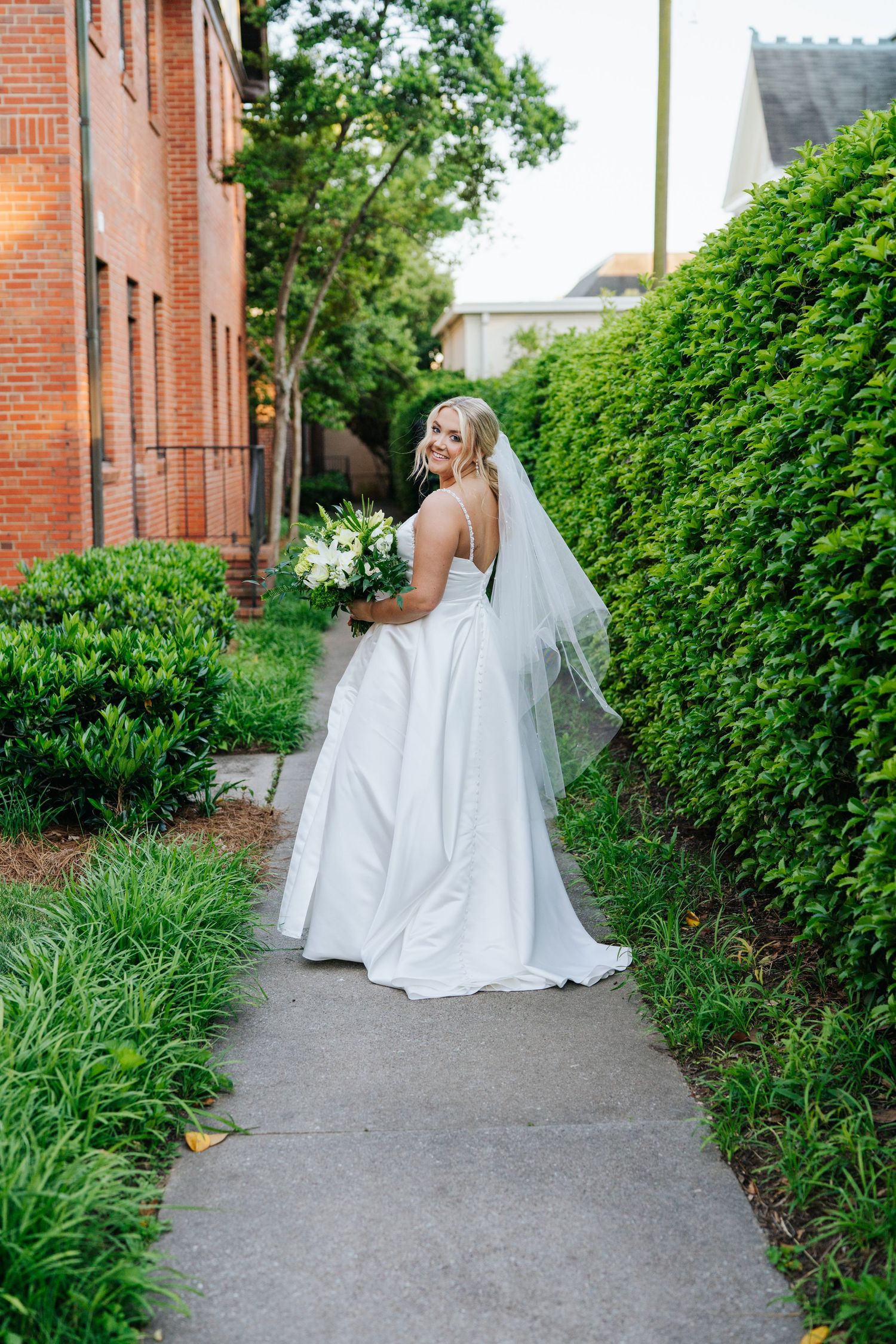 A bride holding a bouquet stands on a garden path between brick buildings and hedges.