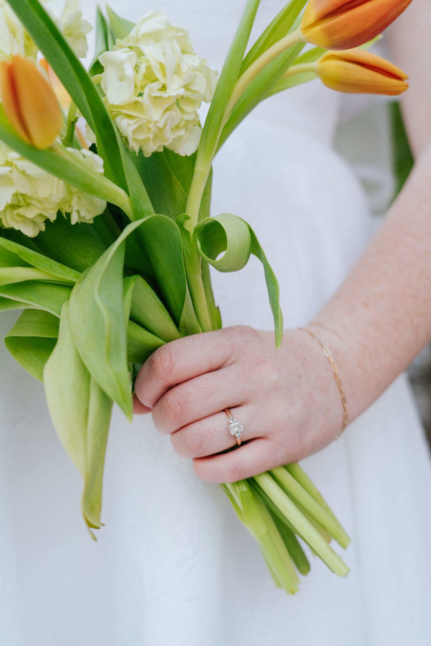 Close-up of hands holding a bouquet of orange tulips and white hydrangeas against a light background.