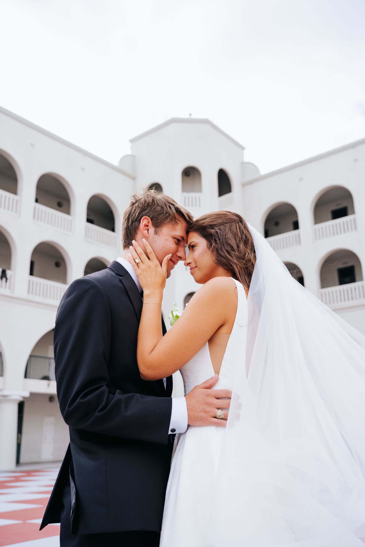 Intimate wedding couple moment captured against white architectural backdrop with flowing veil.