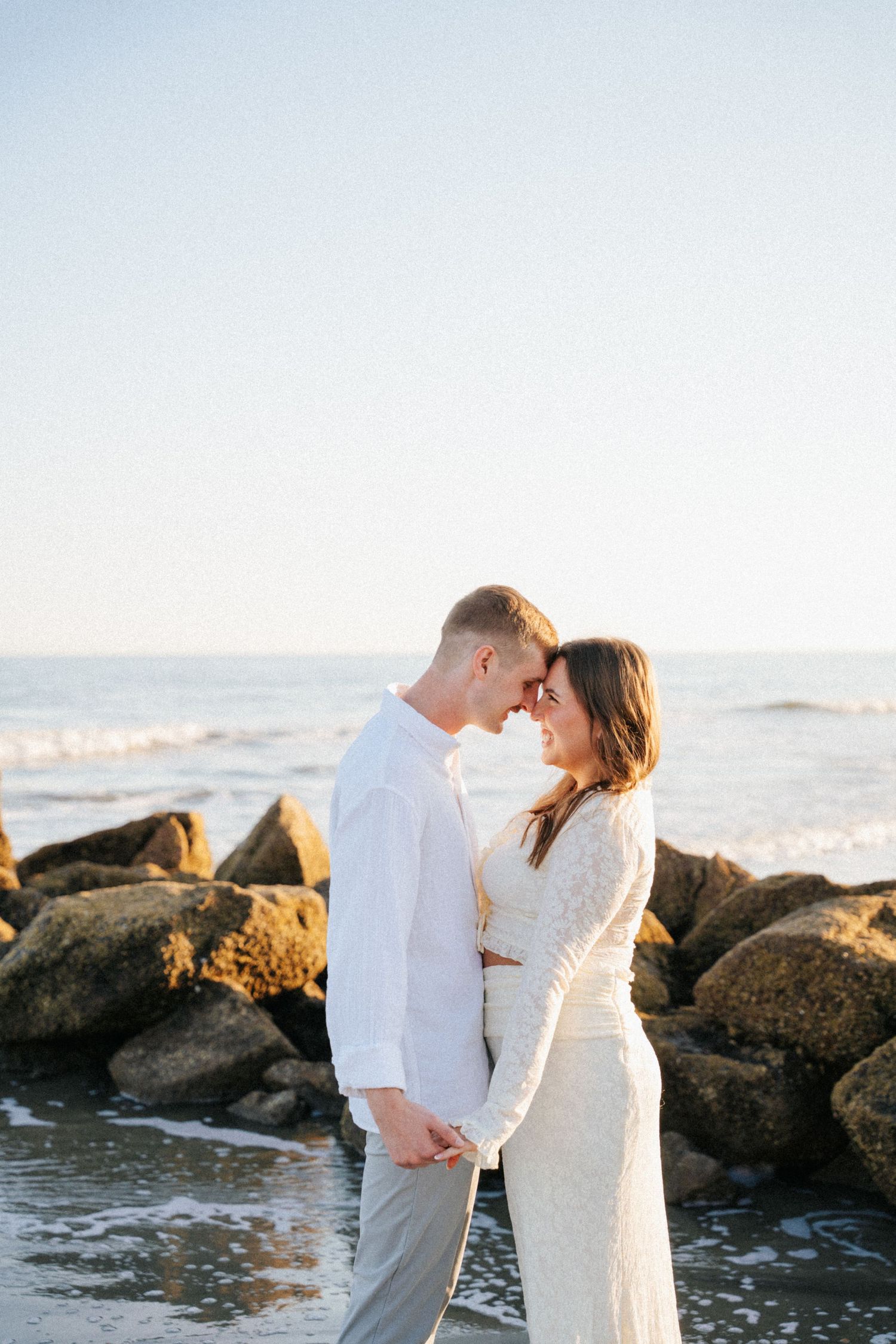 A couple in white attire shares an intimate moment on a rocky beach at sunset with ocean waves in the background.