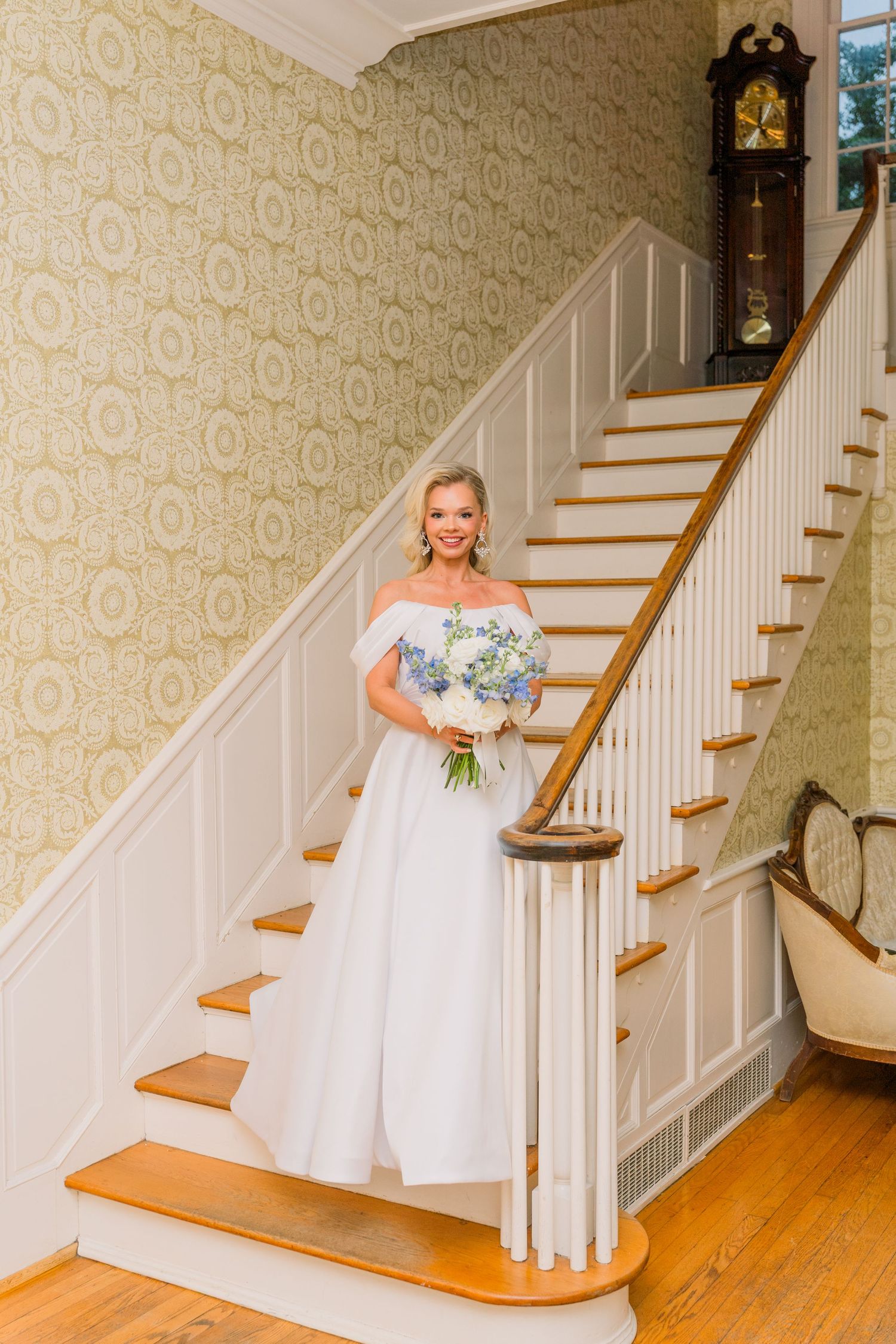 Bride in white off-shoulder gown holding blue bouquet stands on wooden staircase in elegant home with patterned wallpaper.